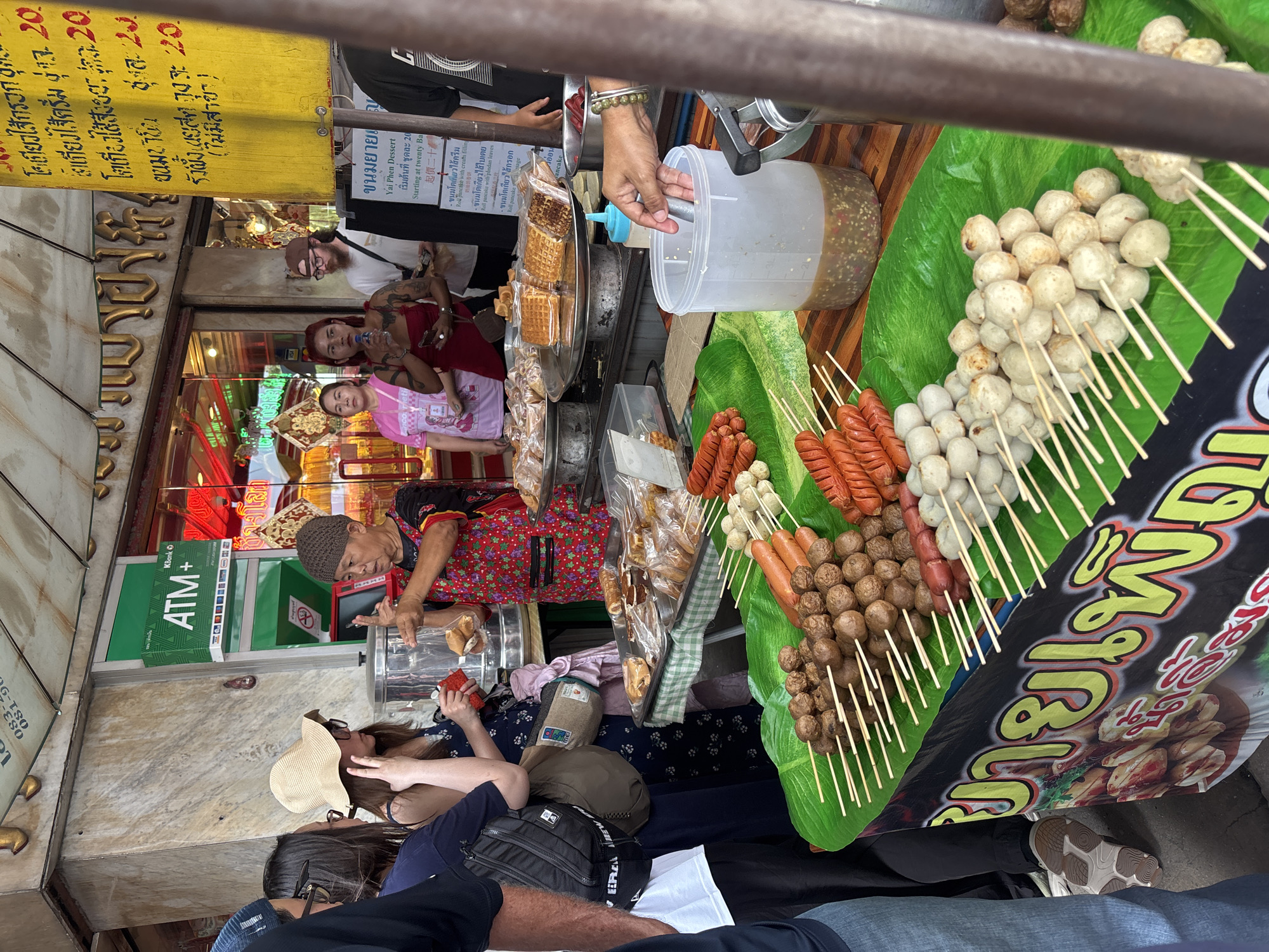 This image depicts a street food stall in Chiang Mai, Thailand, likely within the Chiang Mai City Municipality. The stall is set up outdoors, possibly on a sidewalk or near a market. In the foreground, skewers of various grilled food items are prominently displayed on large green banana leaves. These include numerous small, round meatballs, several skewers of red sausages, and some darker, larger meatballs. To the right, a large clear plastic container holds a dark sauce, and a hand is visible reaching into it with a utensil.
Behind the food display, several people are interacting with the vendor. The vendor, an older woman wearing a patterned apron over a dark shirt and a knitted cap, is seen gesturing with her hands as she appears to be serving or preparing food. She is holding a plastic bag and possibly some bread. In front of the vendor, several customers are gathered, including a woman in a wide-brimmed hat and sunglasses, and another woman in a dark dress. Further in the background, more individuals can be seen, including a man wearing a cap and a woman in a pink t-shirt.
The stall itself has various elements of a commercial operation, including signs with Thai script. One sign, a bright yellow one, displays pricing information and food descriptions. Another sign, green with white lettering, says "ATM+" and appears to be an advertisement for a bank. The environment suggests a bustling commercial area, with the typical sights and sounds of a street market. The overall scene captures a moment of commerce and daily life at a popular food vendor.