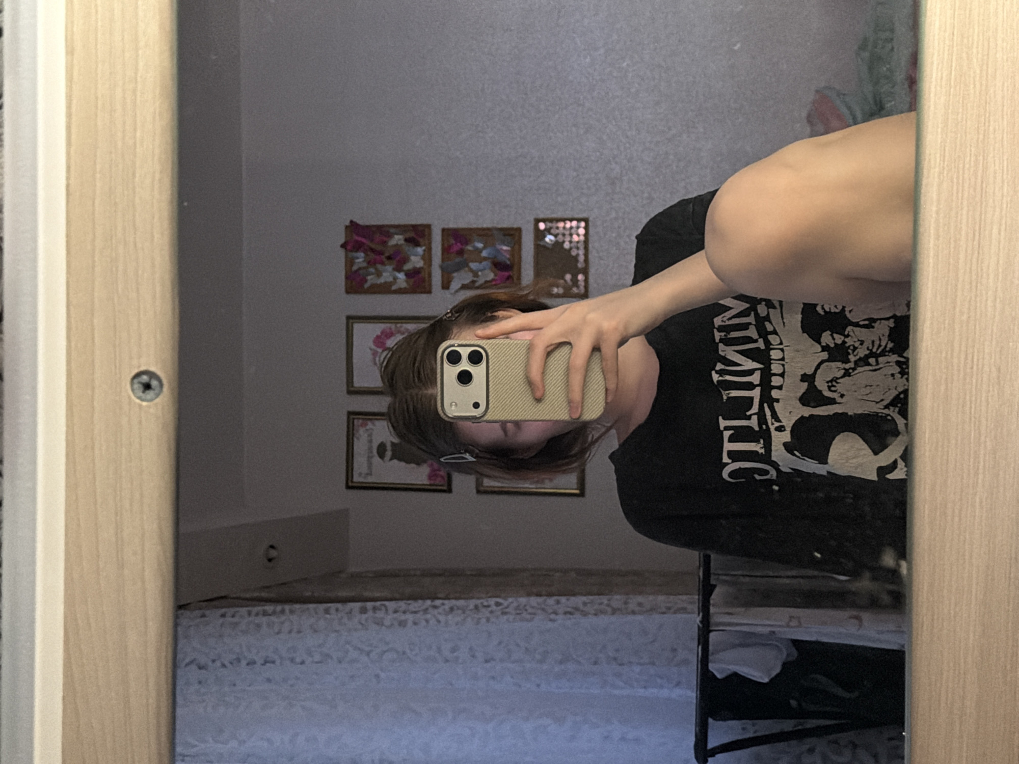 This mirror selfie, taken indoors in Noviy, Russia, captures a person partially obscured by their smartphone. The subject, with short to medium brown hair, holds a light-colored, textured phone case with a triple camera setup, concealing their face. They wear a black t-shirt featuring an inverted, stylized white graphic, with visible letters that appear to read "VINTIC" or similar. A bare arm and a leg are also visible in the reflection, suggesting a casual pose.
The room, as seen in the mirror, has light grey, textured wallpaper. Decorating the wall are several framed art pieces, including two square frames with what look like floral or fabric designs, and another with small reflective squares. Below these, two smaller framed items are also visible. A bed with a white, patterned bedspread can be seen in the lower portion of the reflection. To the right, a black metal shelf or rack with items stored on it is partially visible. The mirror is set within a light-colored wooden frame, with a screw clearly visible on the left side. The lighting is ambient, characteristic of an indoor setting.