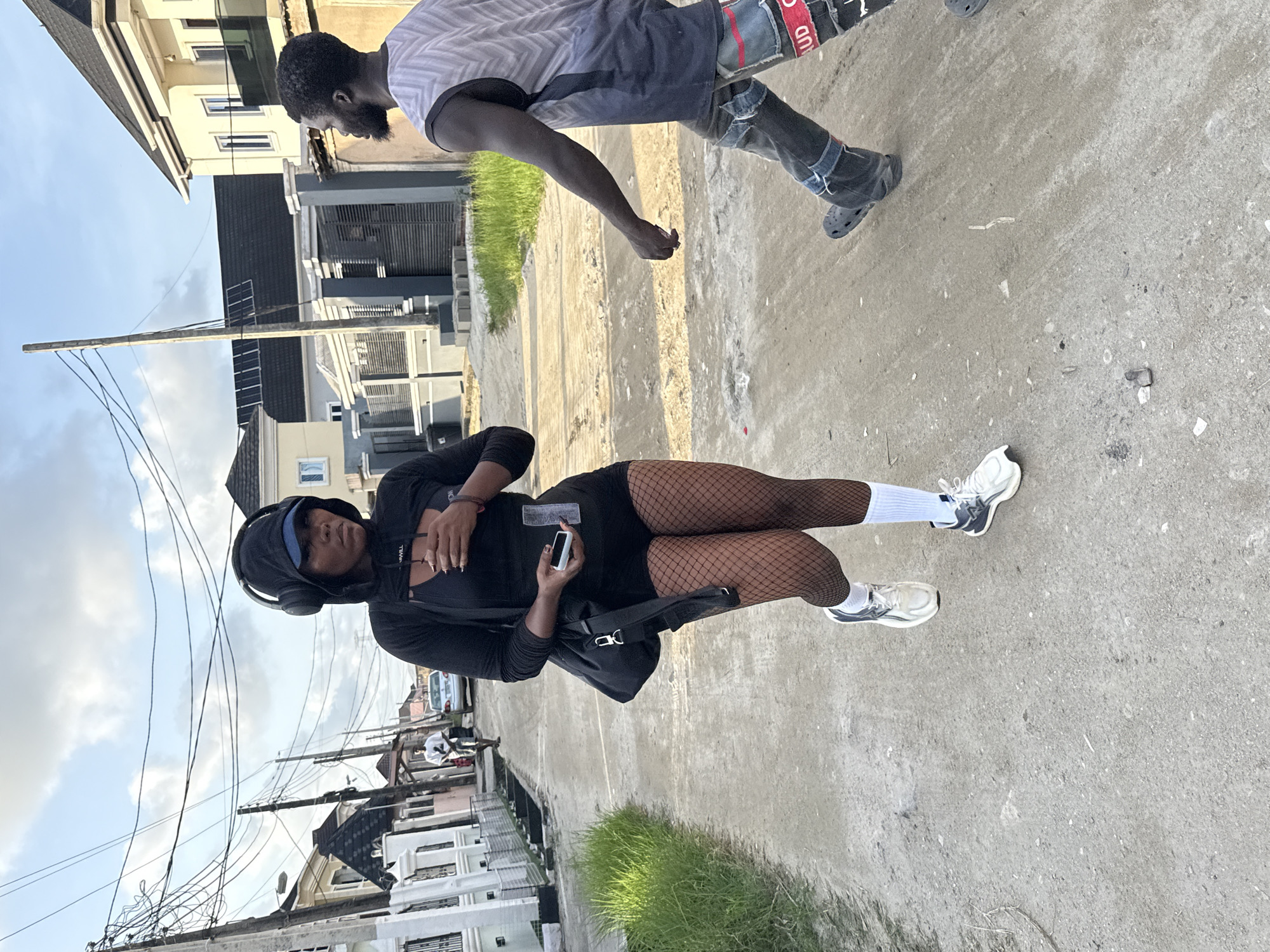A young woman wearing a black hooded jacket, fishnet stockings, white socks, and white sneakers stands on a concrete street in a residential area of Ado, Nigeria. She holds a phone and carries a black shoulder bag. Another person, a Black man in a light grey t-shirt and dark jeans with patchwork, walks nearby. The street is lined with houses, and overhead are numerous power lines attached to utility poles. The sky is partly cloudy.