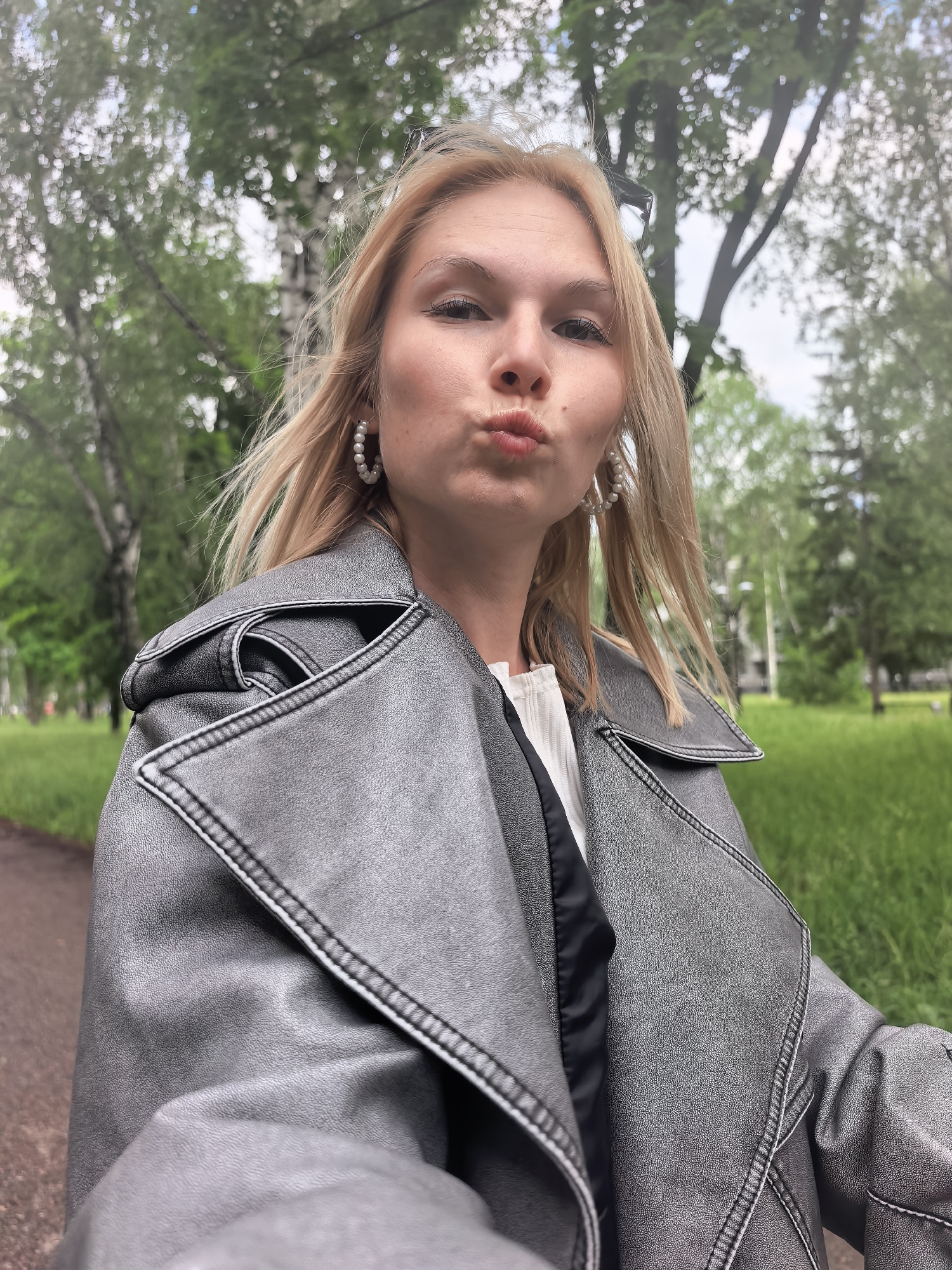 A woman is taking a selfie outdoors. She is wearing a silver/grey jacket, a white shirt, and pearl earrings. Her blonde hair is shoulder-length. She is making a kissy face at the camera. The background is blurred, but it appears to be a park or green space with trees. Based on the metadata, the location is Sterlitamak, Russia.