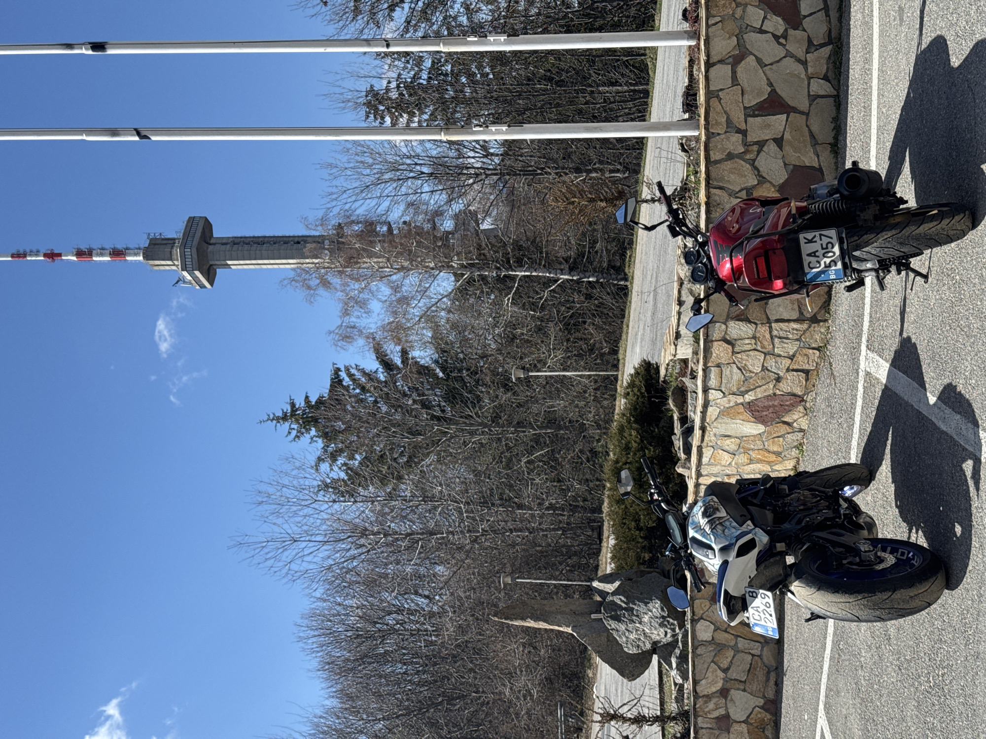 The image depicts two motorcycles parked on an asphalt parking lot in front of a stone wall. In the background, a tall tower, possibly a TV or communications tower with a red and white striped top, stands against a clear blue sky. Bare trees are visible between the motorcycles and the tower. The motorcycles appear to be parked near a scenic viewpoint or tourist location. Based on the tower design, the location may be near Kopitoto lookout point, Vitosha mountain, Sofia, Bulgaria. The motorcycles have license plates. The red motorcycle's license plate reads "CAK 5057", and the blue motorcycle's license plate reads "CA 8269".