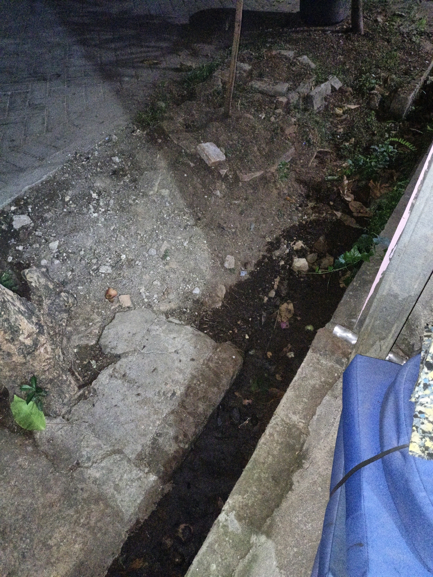 The image shows a narrow drainage ditch filled with dark water and debris. The ditch is lined with concrete on both sides, with one side appearing to be a raised concrete structure. The surrounding area is a mix of dirt, gravel, and some scattered vegetation. There are also some broken concrete pieces and what appears to be a discarded brown object near the edge of the ditch. To the right, a large blue object, possibly a tarp or fabric, is visible, partially obscuring a patterned surface. The lighting suggests it is nighttime or a very dimly lit area. The location is stated to be in the city of Cikal, Indonesia, which is known for its industrial and agricultural activities, and often experiences heavy rainfall, contributing to the presence of drainage systems like the one depicted.