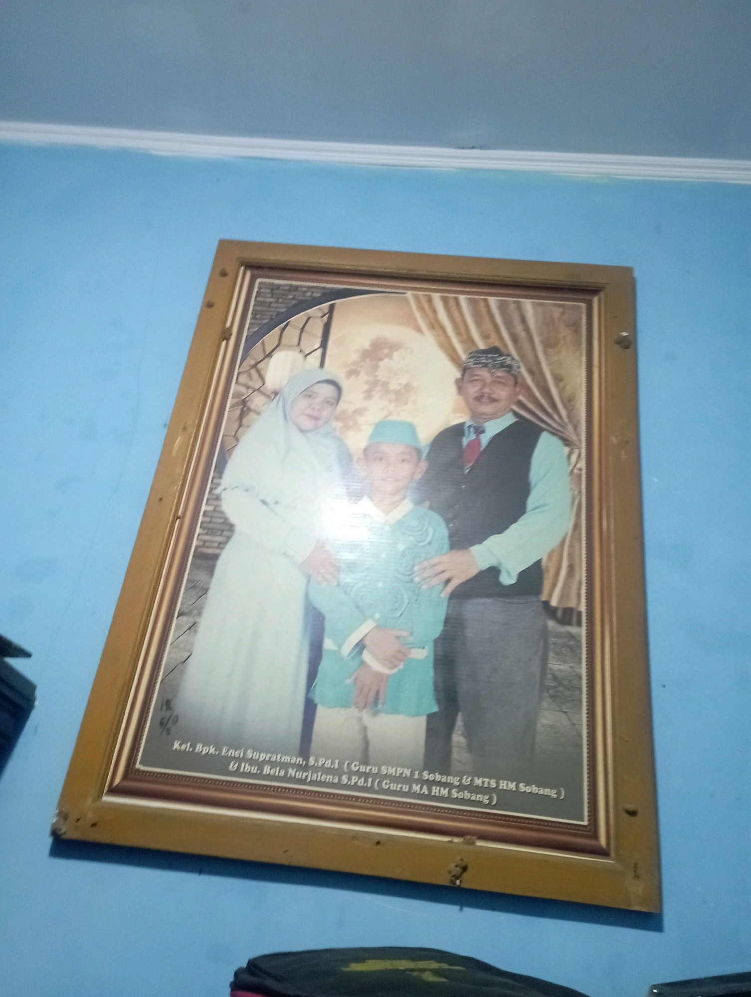 A framed family portrait hangs on a blue wall, angled upwards. The portrait shows a family of three: a woman in a light-colored hijab and dress, a man wearing a traditional patterned cap, a vest, a collared shirt, and a tie, and a young boy in a light blue cap and matching outfit. The family is posing for the photo, with the parents standing and the boy positioned in front of them. Inscriptions are visible at the bottom of the frame, suggesting this is likely a family photo from Senangsari, Indonesia. The overall scene is dimly lit, with the blue wall showing slight discoloration and damage near the bottom edge of the frame.