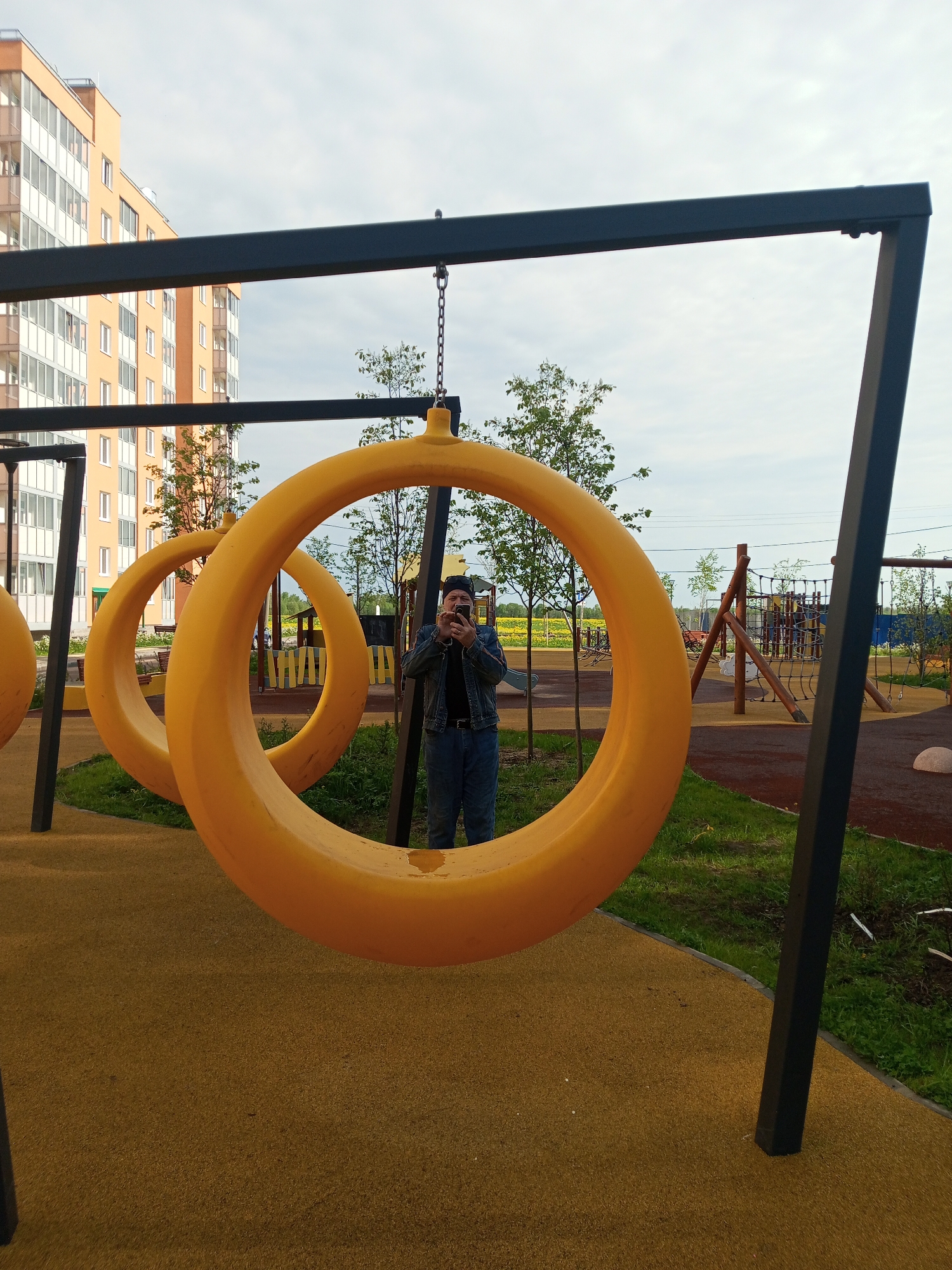The image, taken in the city of Kommunar, Russia, shows a man standing inside a large yellow circular swing at a playground. He is facing the camera and holding a smartphone, presumably taking a picture. The man is wearing a dark jacket and jeans. Two other similar swings are visible nearby. In the background, there is a multi-story building, trees, and other playground equipment. The ground is covered in a soft, rubbery material. The sky is overcast.