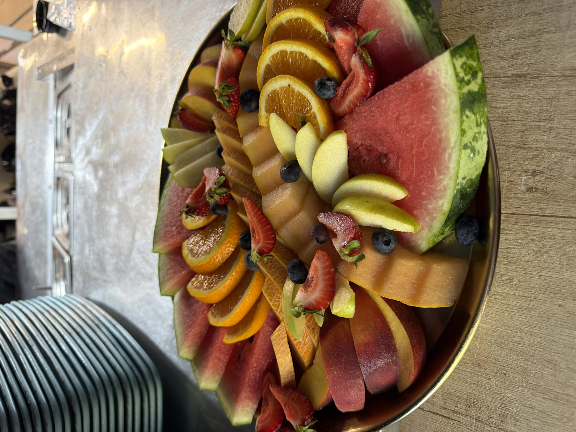 A platter of sliced fruits, including watermelon, oranges, peaches, cantaloupe, and strawberries, is arranged on a metallic surface, likely a countertop or table. Several blueberries are scattered among the fruits. In the background, a stack of light blue plates is visible, along with a metallic counter and some indistinct figures. The overall setting suggests a food service area, possibly a catering event or buffet, in Gorna Malina, Bulgaria.