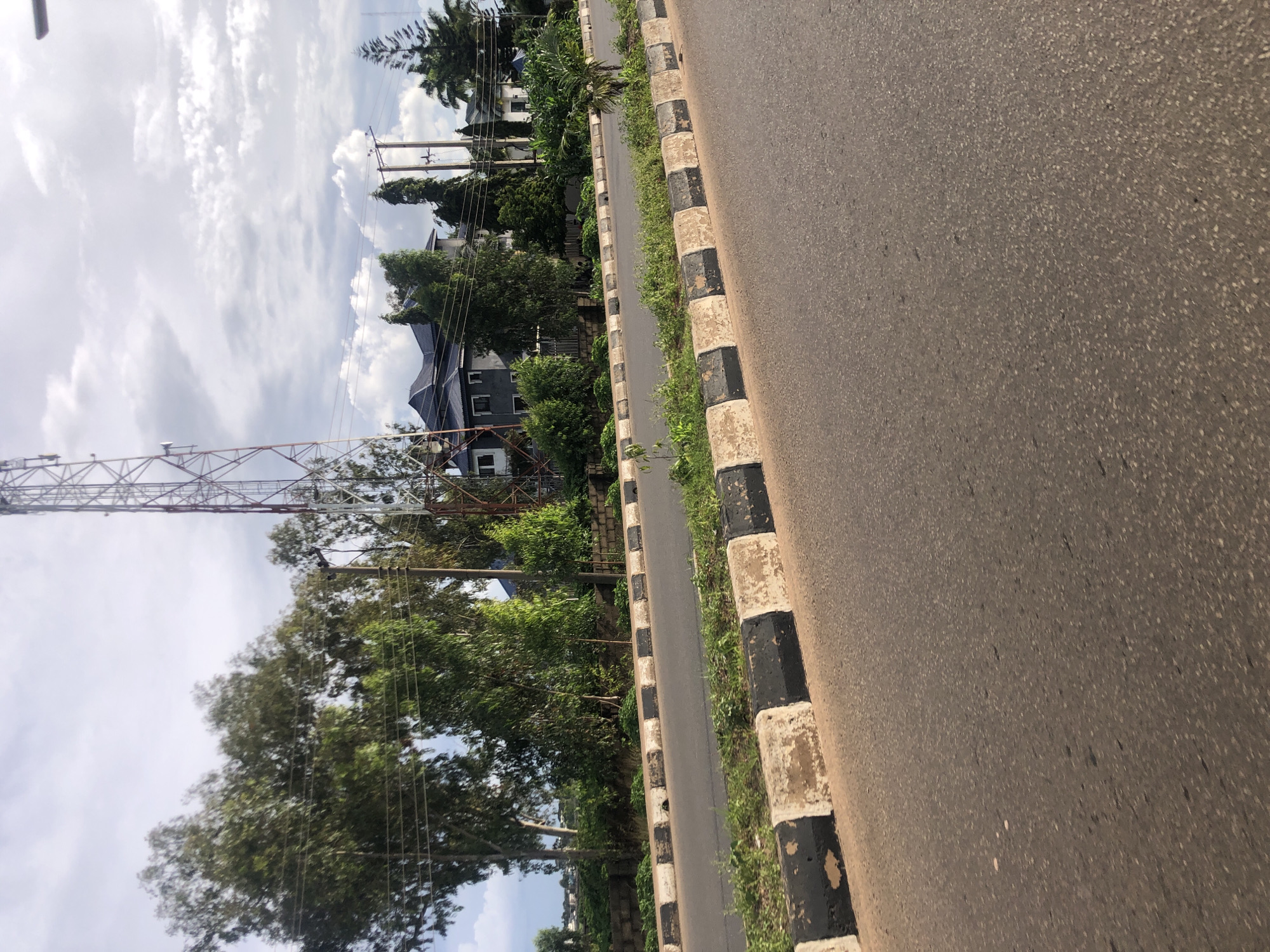 A road in Jos, Nigeria, curves to the right, bordered by a black and white striped curb. Lush green trees line the side of the road, behind which a large, dark-roofed building stands. A tall, red and white telecommunications tower looms in the background. The sky is filled with a mix of clouds and blue sky, suggesting it is daytime.