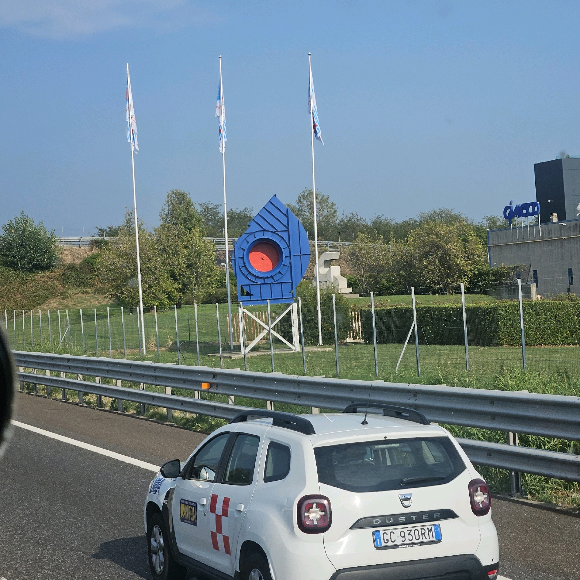 The image captures a view from a vehicle on a highway in Trezzano Rosa, Italy. In the foreground, a white Dacia Duster with an Italian license plate "GC 930RM" is visible on the right lane. The car has a company logo on its side, partially obscured. A metallic guardrail runs along the edge of the highway.
In the middle ground, a large, abstract blue sculpture with a red circular element is displayed on a grassy area. This sculpture is supported by a white X-shaped structure. Behind the sculpture, there are trees and a building with a prominent blue logo that appears to spell "CHICCO". Two flags are also flying on flagpoles near the sculpture. The environment is characterized by a clear blue sky and green vegetation. The overall scene suggests an industrial or commercial area adjacent to a highway.