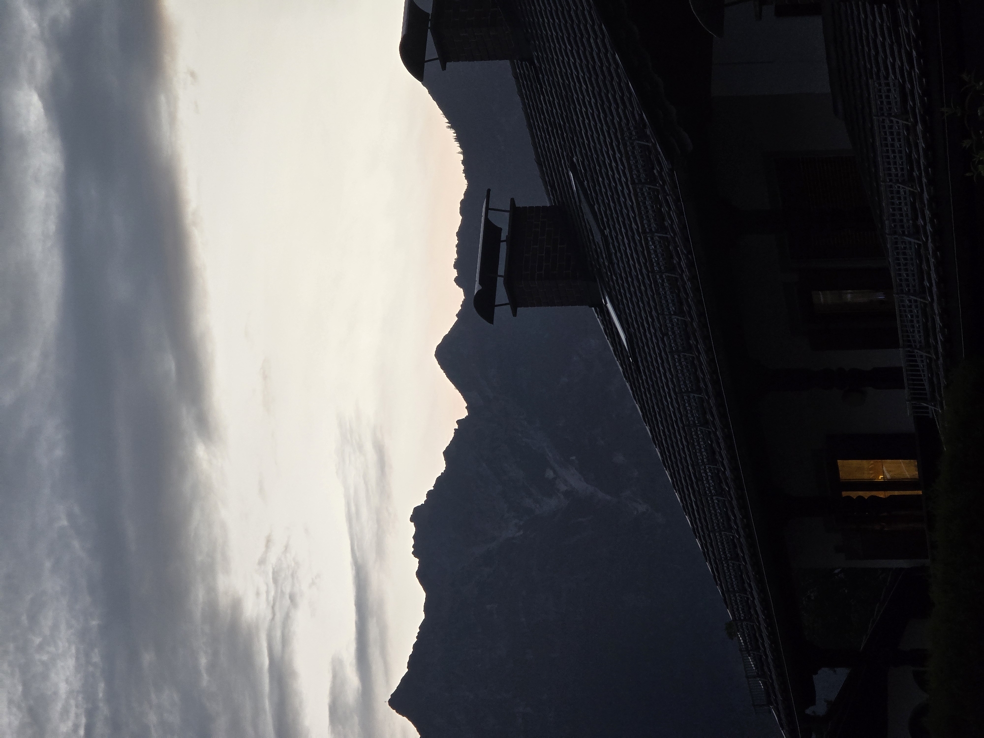 The image depicts a silhouetted scene of a house and mountains at dusk, likely in or near Garmisch-Partenkirchen, Germany. The foreground is dominated by the dark silhouette of a tiled roof with two brick chimneys. The roof slopes downwards from right to left, with the left portion of the house receding into darkness. One window on the lower level of the house glows with a warm, orange light. Beyond the house, a range of dark, rugged mountains fills the middle ground, their peaks silhouetted against a pale, overcast sky. The sky transitions from a lighter hue at the horizon to darker, cloud-laden tones towards the top of the frame. The overall atmosphere is subdued and serene, characteristic of twilight in a mountainous region. There are no people or animals visible in the image.