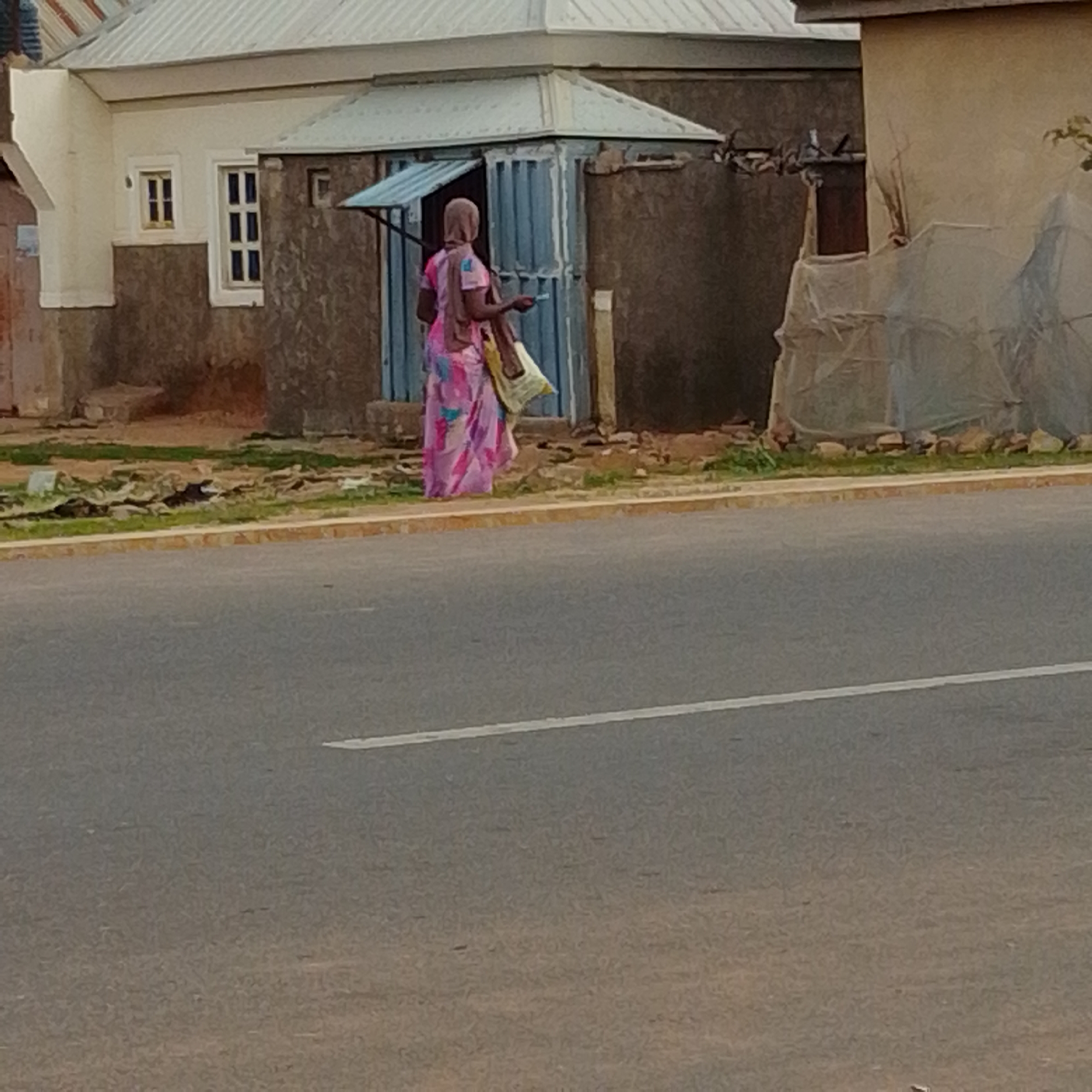 A woman in a pink and blue patterned dress and a light brown headscarf walks past a building on a dusty street. She is carrying a light-colored bag over her shoulder and a blue object in her right hand. The building behind her has a light-colored facade, a corrugated metal roof, and a blue metal door. There is a patch of green grass and debris in front of the building, separating it from a paved road with a white line. The time of day appears to be daytime, and the weather seems clear. The scene suggests a casual street setting in an urban or semi-urban area. There is no visible text in the image.