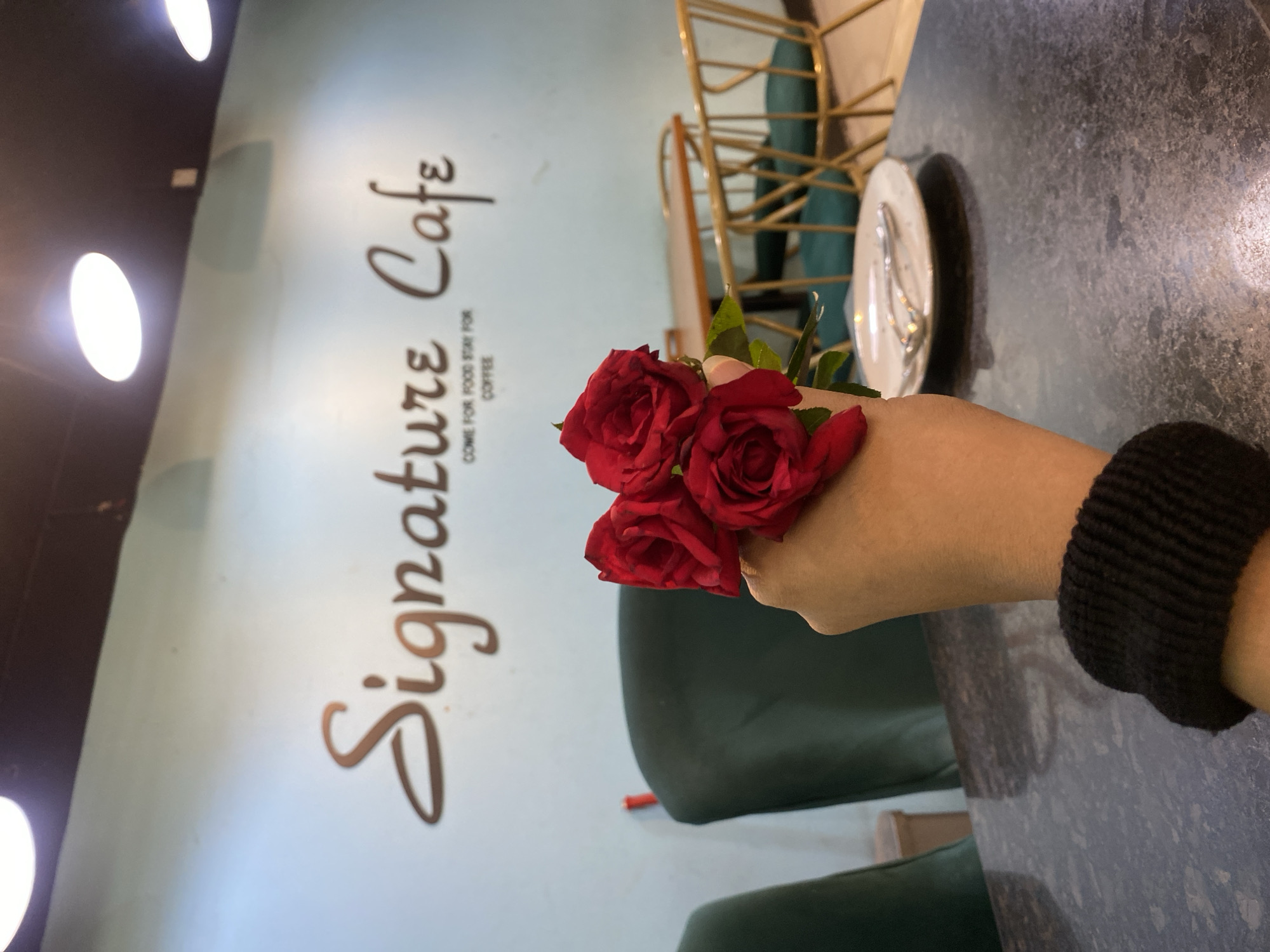 A close-up shot focuses on a hand holding three red roses in the foreground, with a dark-speckled table surface visible below. In the mid-ground, a plate with a spoon and fork sits on a table. Beyond that, dark green chairs are arranged around tables. The background features a light blue wall with "Signature Cafe" inscribed in a cursive font and smaller text below reading "COME FOR FOOD STAY FOR COFFEE". The cafe setting suggests it is likely located in a city like Dhaka, Bangladesh, given the typical cafe ambiance in such urban environments. The lighting appears to be from overhead fixtures, possibly spotlights, indicated by bright circular areas on the ceiling.