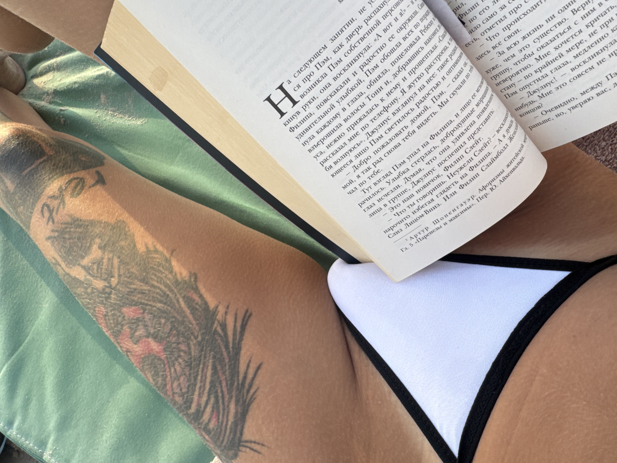 A close-up, overhead shot shows a person lying down, reading a book. The subject is wearing a white bikini with black trim. Their left arm, which has a large tattoo of a feather and text, is resting on a green fabric, possibly a towel or blanket. The book is open, and text is visible on the pages. The lighting suggests it is daytime, possibly outdoors given the natural light. The mood appears relaxed and focused on reading. Visible text on the book includes Russian words, some of which appear to be literary or philosophical titles and authors like "Артур Шопенгауэр". The text also includes chapter numbers and possibly translation credits. The scene is intimate and personal, focusing on the act of reading in a relaxed setting.