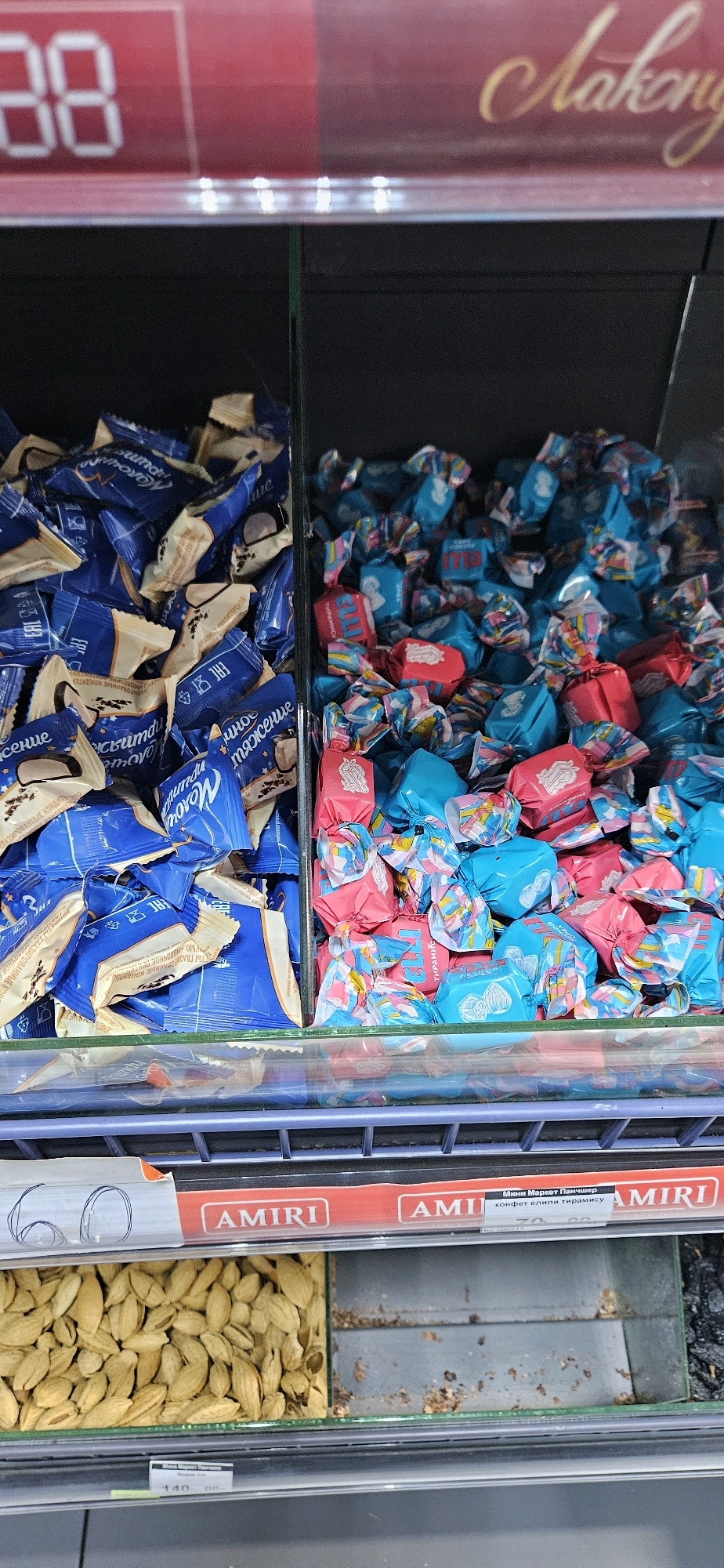 The image shows a close-up, eye-level view of a refrigerated display case in a store. The display case is divided into two sections, with candies on the left and pink and blue wrapped candies on the right. The left section contains blue and white wrapped candies, with some visible text in Russian. The right section has a chaotic assortment of pink, blue, and multi-colored wrapped candies. Below the candies, there is a shelf displaying what appears to be dried fruit or nuts on the left, and an empty section on the right. The overall environment suggests a retail setting, likely a grocery store or a confectionery shop. The red signage at the top of the display has some Cyrillic text and a numerical value. The image does not provide any specific geographical markers that would indicate its location in Dushanbe, Tajikistan, or any other city. There are no people or animals visible in the frame.
