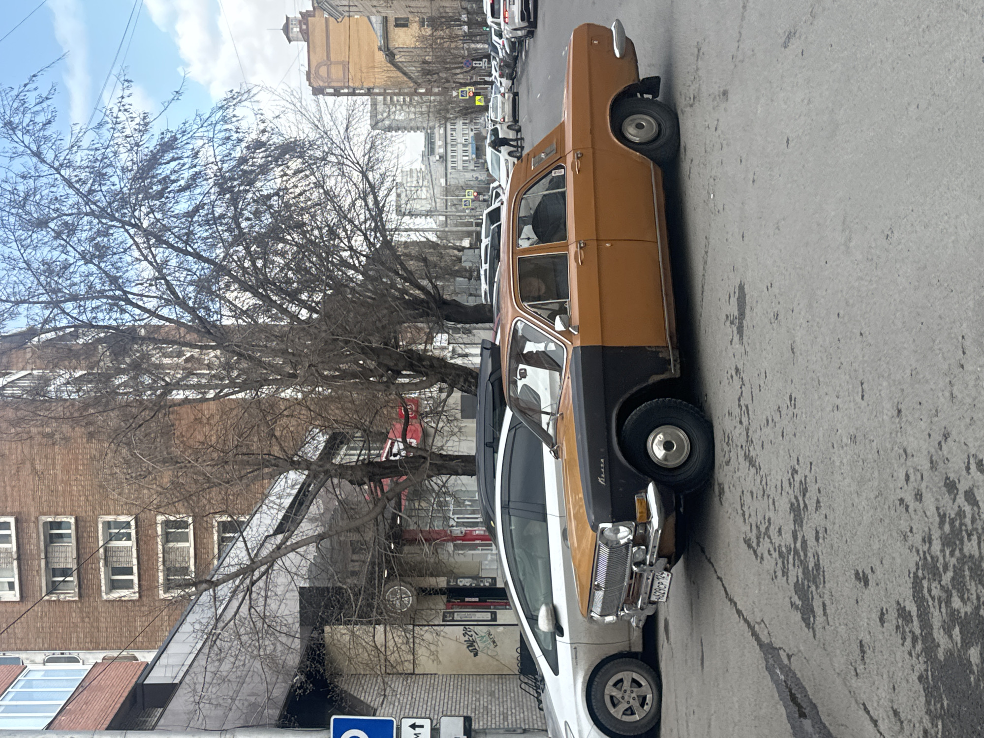 The image depicts a street scene in Krasnoyarsk, Russia. A two-tone, orange-and-black vintage car is parked on the side of the road, next to a white car. In the background, a row of parked cars is visible. Buildings line the street, and a bare tree stands beside the vintage car. Pedestrian crosswalks are marked on the road. The sky is partly cloudy.