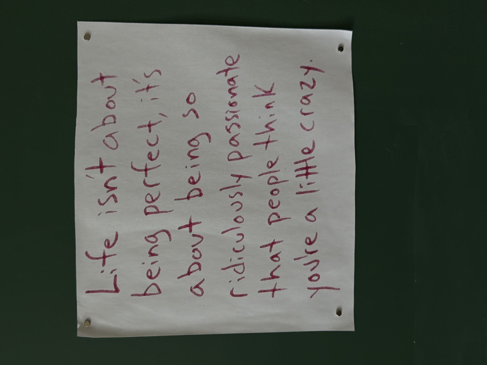 A rectangular sheet of white paper, with a few small holes punched near the corners and attached to a dark green surface, is vertically oriented in the image. Written in burgundy marker on the paper is the text, “Life isn’t about being perfect, it’s about being so ridiculously passionate that people think you’re a little crazy.” The paper appears to be held up by tacks in the holes near the corners. The location is in Unknown, Unknown.