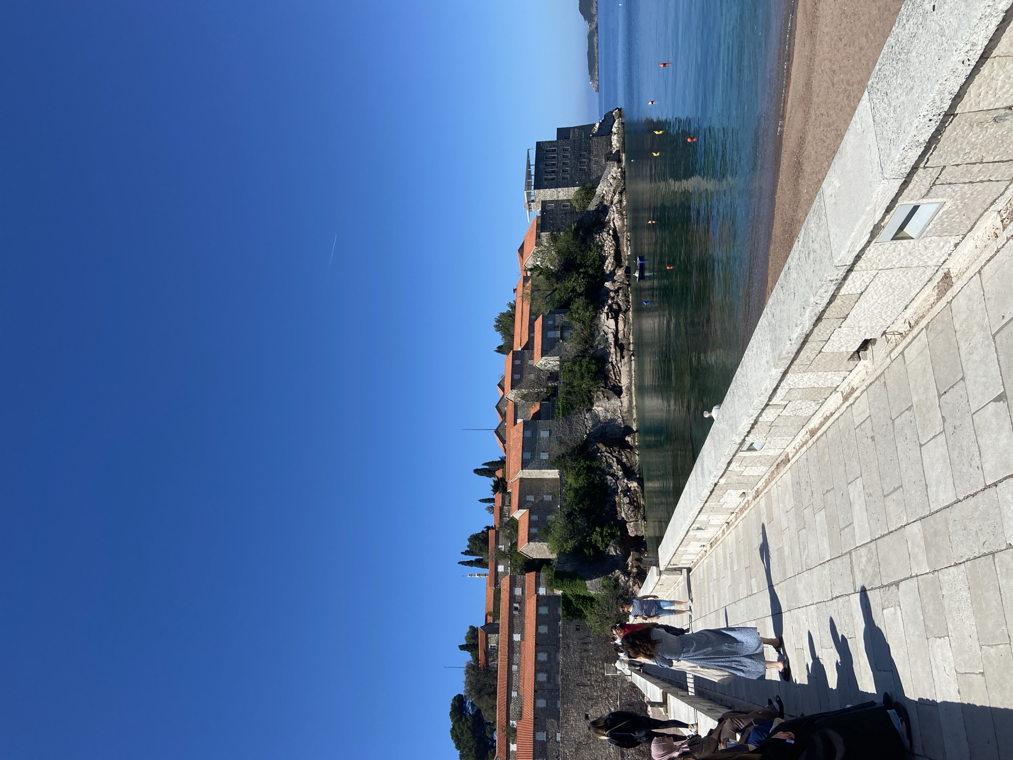 The image shows a view in Sveti Stefan, Montenegro, taken from a paved walkway looking towards the island resort. To the left, the clear blue sky takes up a large portion of the frame. The island resort, composed of buildings with red-tiled roofs, stands on a rocky outcrop, bordered by trees and vegetation. The water reflects the buildings and sky. In the foreground, the walkway is visible with people walking along it, their shadows cast on the pavement. The sea is on the right side of the frame with a sandy beach visible in the distance. Floating buoys of various colors are visible in the water.