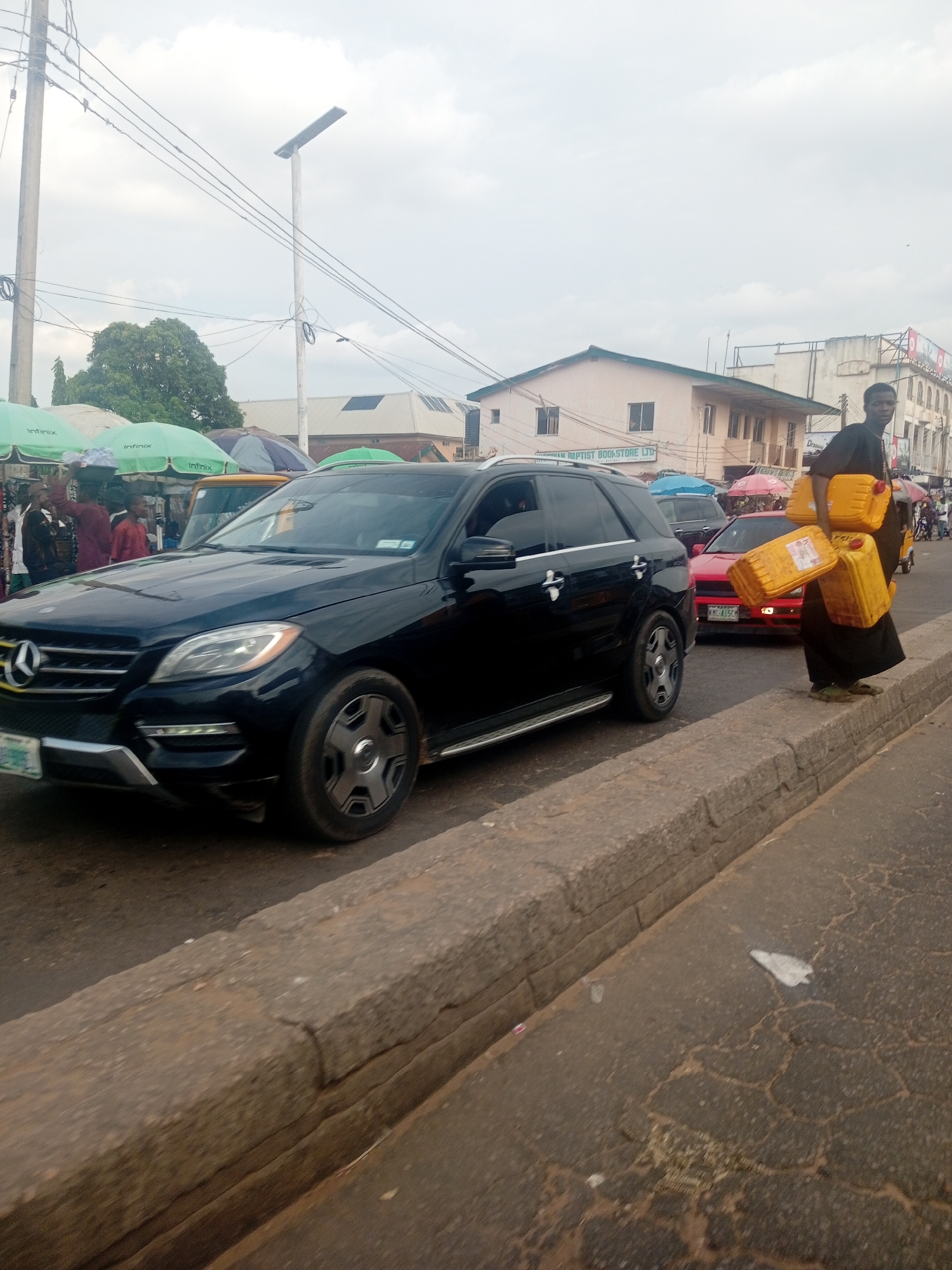 The image captures a street scene in Maiburiji, Nigeria, likely during daylight hours under an overcast sky. The foreground is dominated by a black Mercedes-Benz SUV, suggesting a busy urban environment. To the right, a person, dressed in dark clothing, is walking along a raised concrete curb, carrying several yellow jerrycans, which appear to be filled with liquid, possibly fuel or water. The jerrycans are stacked and balanced, indicating a task of transport or distribution.
Behind the SUV and the individual, several other vehicles are visible, including a red car and what appears to be a yellow van or truck. The background reveals a row of buildings, one of which has signage indicating "CHURCH AND BAPTIST BOOKSTORE LTD." This detail provides a specific clue to the location. Several colorful market umbrellas are set up, suggesting a marketplace or a roadside vendor area, with people mingling and interacting. The presence of utility poles and overhead wires further emphasizes the urban setting.
The overall atmosphere seems to be one of commerce and daily life unfolding in a bustling environment. The expressions of the people are not clearly discernible due to the distance and image quality, but the activity of carrying the jerrycans suggests a purpose-driven movement. The concrete curb the person is walking on is weathered and cracked, and the road surface shows signs of wear. The presence of multiple vehicles and pedestrians implies ongoing traffic and activity.