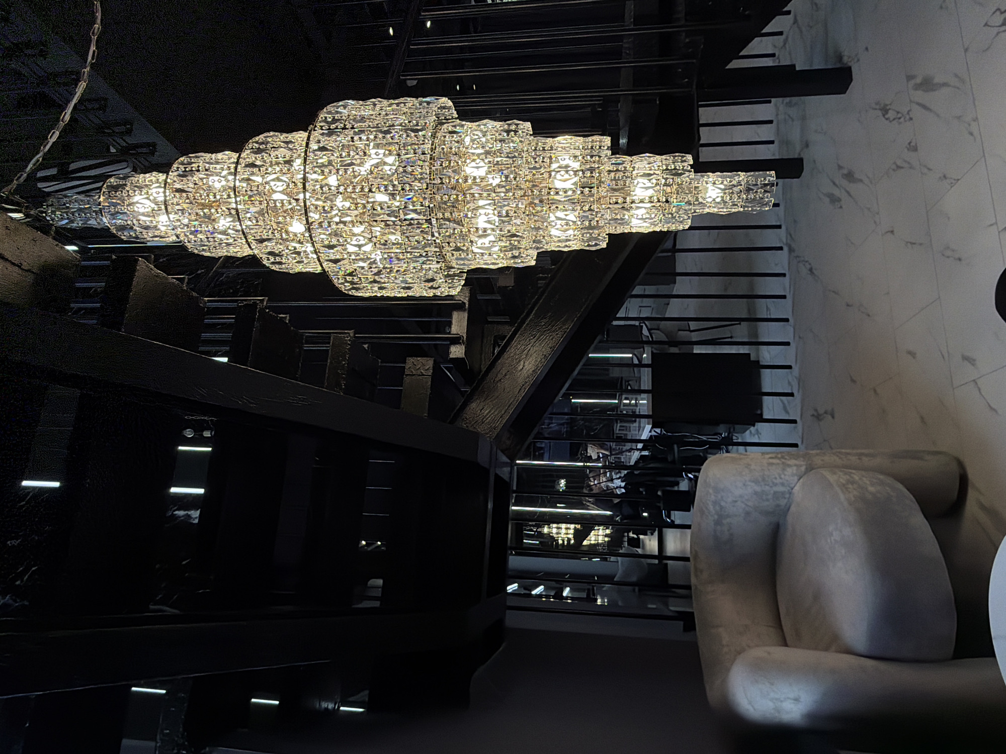 A low-angle interior shot captures a dimly lit space with a prominent, multi-tiered crystal chandelier hanging from the ceiling. The chandelier features several cascading layers of faceted crystals, reflecting and refracting the warm light emanating from within. To the left of the chandelier, a plush, light-colored armchair with rounded arms and a circular seat is visible. The armchair is positioned on a floor with a marble-like pattern. Behind the armchair and to the right, a dark, industrial-style staircase with metal railings ascends. The staircase appears to be constructed from black metal beams and treads. In the background, through the open structure of the staircase and beyond a railing, glimpses of other areas of the establishment are visible, including tables, chairs, and more vertical lighting fixtures. The overall ambiance suggests a modern, somewhat luxurious, and possibly dimly lit establishment. There are no explicit details that would indicate the location is in Укромновское сельское поселение, Ukraine.
