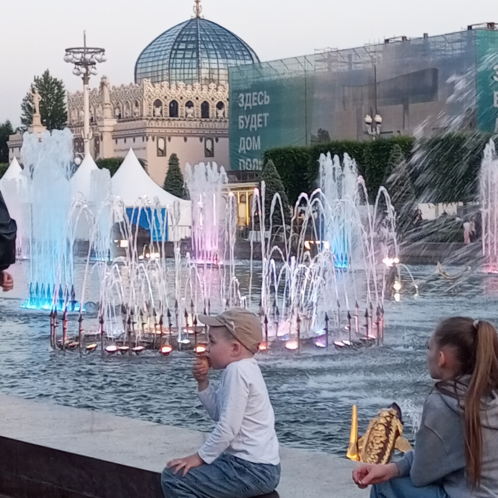 This image captures a public park scene at dusk or night, identified as the Exhibition of Achievements of National Economy (VDNKh) in Moscow, Russia.
In the background, a prominent building with a large glass dome, characteristic of the "Space Pavilion," dominates the skyline. To its left, an ornate, light-colored building with classical architectural details and statues is visible. To the right of the domed structure, a building under construction or renovation is obscured by green scaffolding netting. Russian text printed on the netting reads, "ЗДЕСЬ БУДЕТ ДОМ" (HERE WILL BE THE HOUSE).
The midground is dominated by multiple fountains actively spraying water into a large basin. These fountains are illuminated with various colored lights, including blue, pink, and orange, creating vibrant reflections on the water's surface. White conical tent structures are visible behind the fountains on the left side, and dense green hedges border the fountain area. A tall, multi-lamp street light stands between the fountains and the ornate building.
In the foreground, two children are seated on a low concrete ledge overlooking the fountains. On the left, a young boy, wearing a beige baseball cap, a long-sleeved white shirt, and blue jeans, is positioned in profile, eating from an ice cream cone. To his right, a girl with light brown hair tied in a ponytail, dressed in a grey hooded sweatshirt and blue jeans, sits looking towards the right edge of the frame. A golden, ornate object rests on the ledge near her right hand. The partial arm and shoulder of an adult, clad in a dark jacket, are visible on the far left. The water in the fountain basin shows ripples from the spraying water.