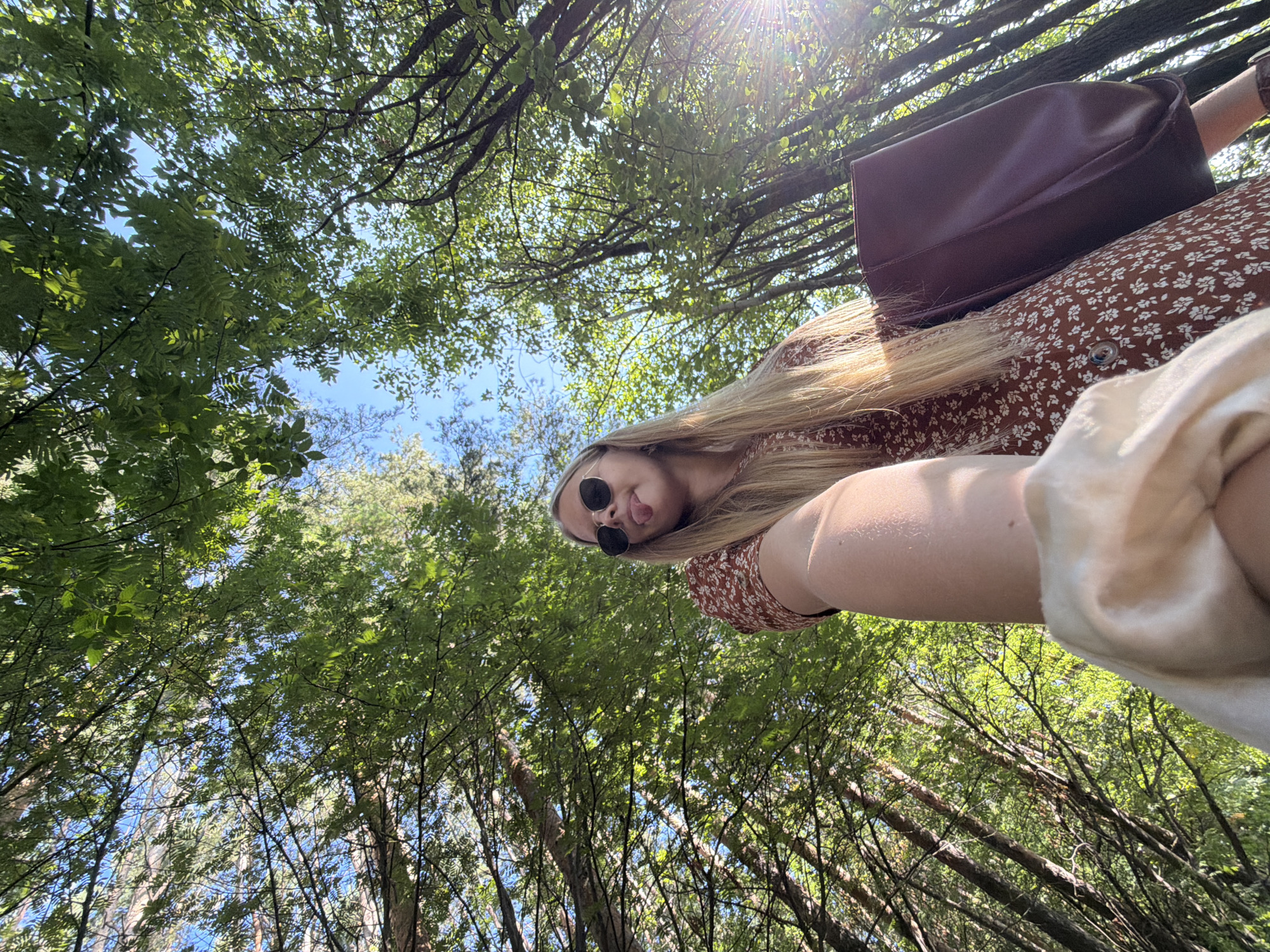 A low-angle, wide shot captures a young woman in sunglasses and a floral print dress holding a dark-colored purse while standing in a forest. The woman's tongue is sticking out, and she is looking towards the camera. The scene is illuminated by sunlight filtering through the dense green canopy of trees, with the bright blue sky visible above. The image is taken from the perspective of someone holding up a phone to take a selfie. The location is not specified to be in the city of Izhevsk, Russia.