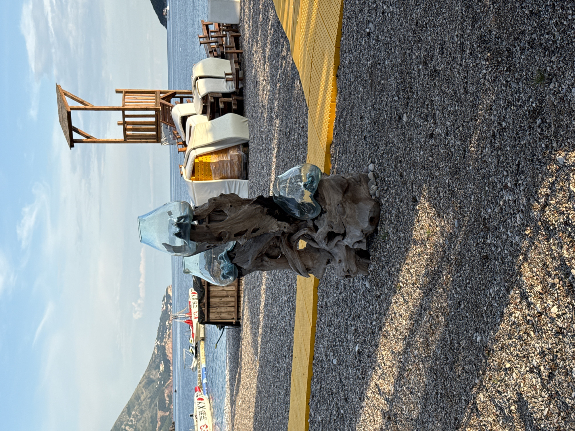 The image captures a scene on a beach near Budva, Montenegro, displaying a blend of natural and constructed elements. In the foreground, a driftwood sculpture topped with three clear glass vessels sits prominently. Adjacent to the sculpture, a bright yellow wooden walkway stretches into the frame.
Behind the sculpture and the walkway, the beach transitions into gravel, giving way to the Adriatic Sea. There are several white beach chairs stacked and ready to be used. An open wooden structure reminiscent of a lifeguard tower stands out near the chairs. In the distance, along the horizon, a rugged, green mountainous landscape extends along the coastline.
In the background on the water, there is a white boat with writing in a non-English language. The sky above is clear with light clouds. Overall, the setting conveys a tranquil seaside atmosphere.