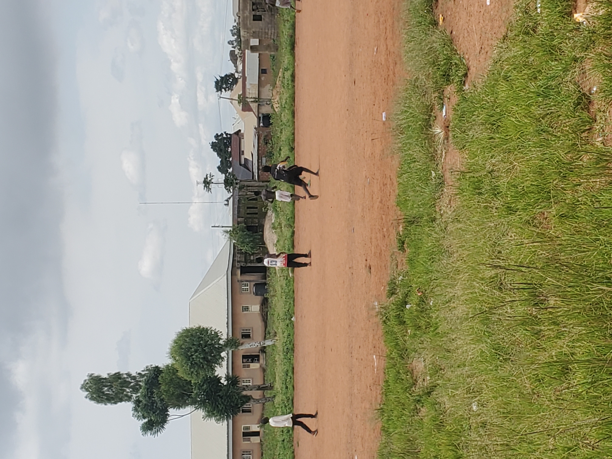 The image captures a group of young men playing soccer on a dirt field in Jos, Nigeria. In the background, a row of residential buildings with light-colored roofs and a few trees are visible. The sky is overcast with clouds. One man in the foreground is wearing a white long-sleeved shirt and dark pants, while another is wearing a white shirt with the number 13 on the back and dark pants. Two other individuals are further down the field, one in dark shorts and a black shirt with yellow accents, and another in light-colored shorts. The field is predominantly dirt with patches of green grass, and there are scattered pieces of debris on the ground.