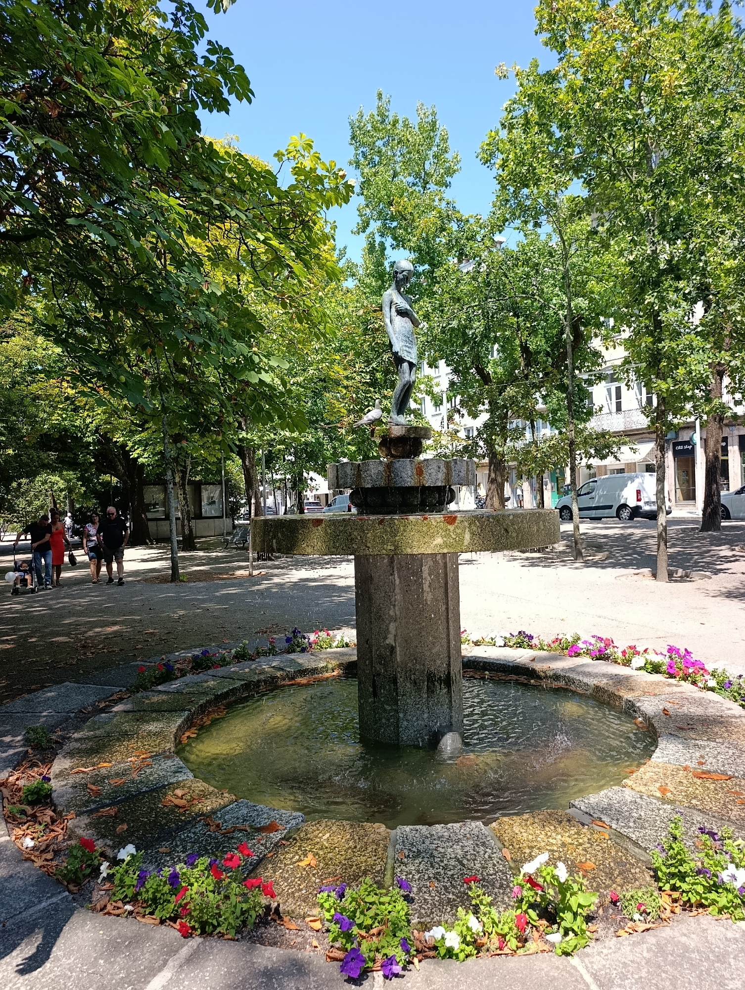 This is a medium shot of a stone fountain in a park in Guimarães, Portugal. The fountain features a tiered basin topped with a statue of a young girl. The lower basin is circular and lined with stone, with colorful flowers planted around its edge. Water trickles from the upper tier into the lower basin. Behind the fountain, several people are walking along a path lined with trees. In the background, there are buildings and a white van parked on the street. The sky is clear and blue.