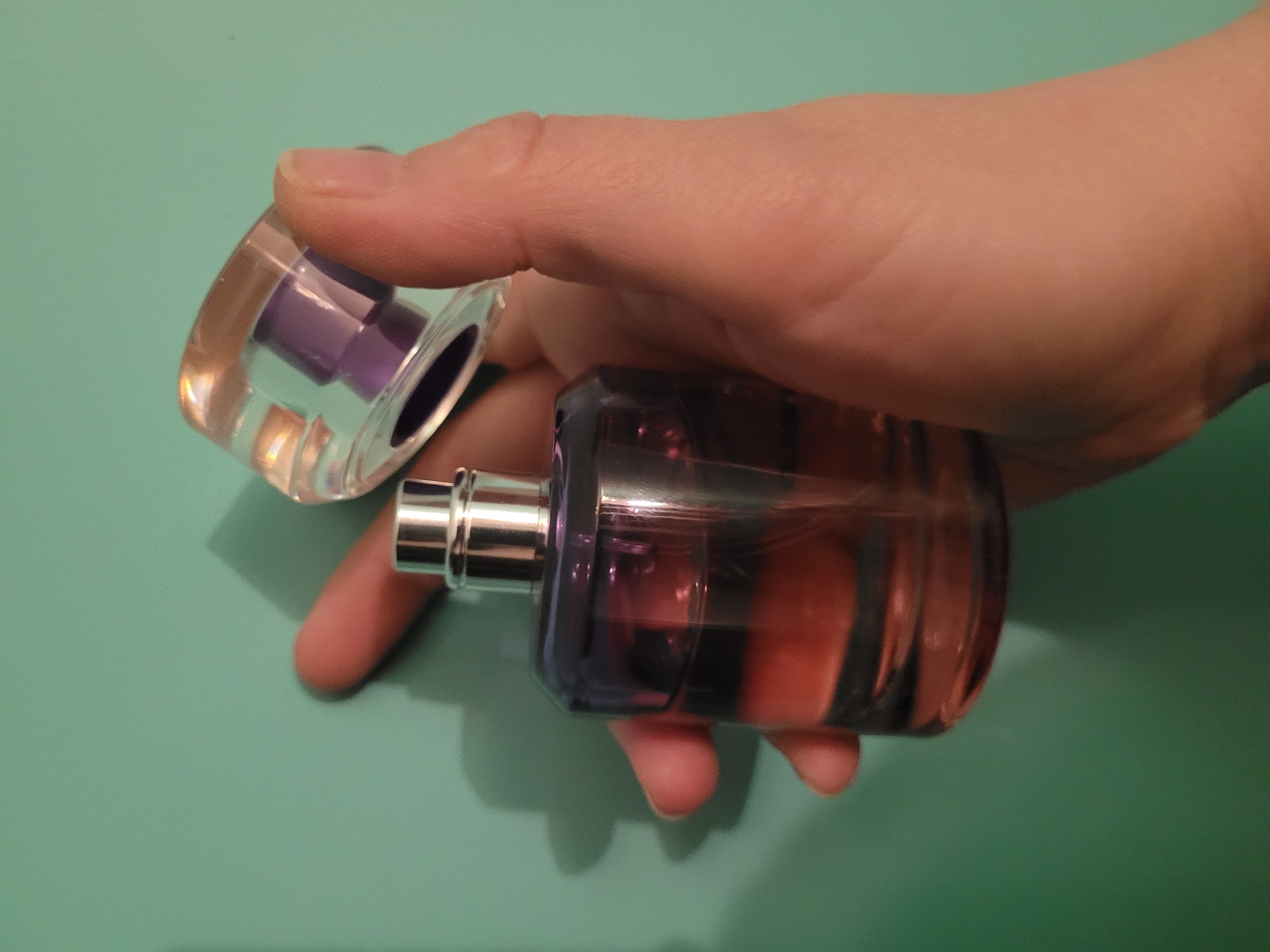 A hand holds an open perfume bottle with a clear cap and silver spray nozzle against a green background. The bottle has dark and light stripes with a purplish hue. The background is a uniform green surface. The image provides no data to support the inclusion of Izhevsk, Russia.