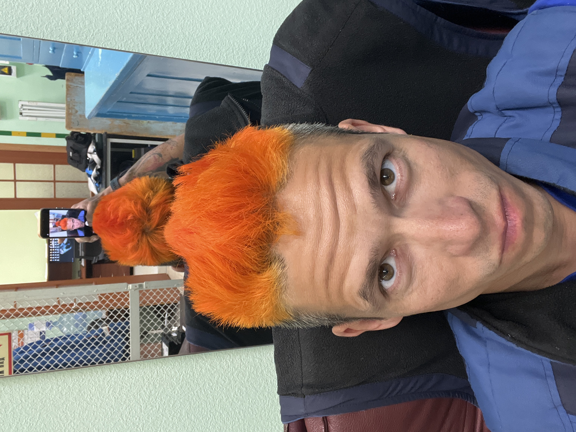 A close-up, angled shot shows a man with short, vibrant orange hair standing in front of a mirror. His expression is slightly quirky, with his eyes looking towards the camera. He wears a blue and black jacket. The mirror reflects the man, holding a phone to take the picture, plus additional elements of the room, including a light blue table-like structure, a metal grid-like rack, and a sign with Russian text. This image was taken in the city of Chaikovsky, Russia.