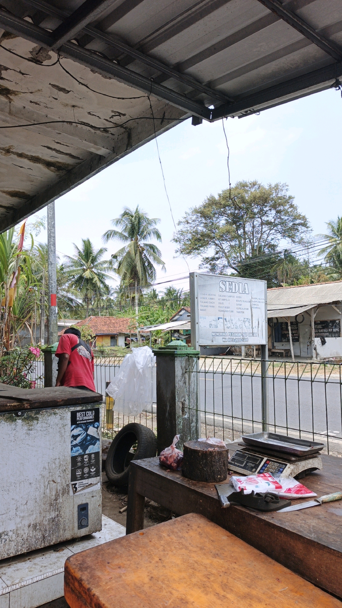 An exterior, eye-level, vertical shot captures a rustic scene in Cibenda, Indonesia, likely a butcher shop. In the foreground, a weathered wooden table displays a meat cleaver, a small knife, a scale, a cylindrical wooden block, and a plastic bag filled with raw meat. A partially visible old chest freezer stands to the left. A person wearing a red shirt is bent over, partially obscured by the freezer and a fence. Behind the fence, a road is visible, with a white sign board that reads "SEDIA" and lists various meat products. The background features houses with red tile roofs, lush green vegetation, and tall palm trees, indicating a tropical or subtropical environment. The sky is clear and bright, suggesting daytime. The overall impression is one of a rural marketplace or food stall.