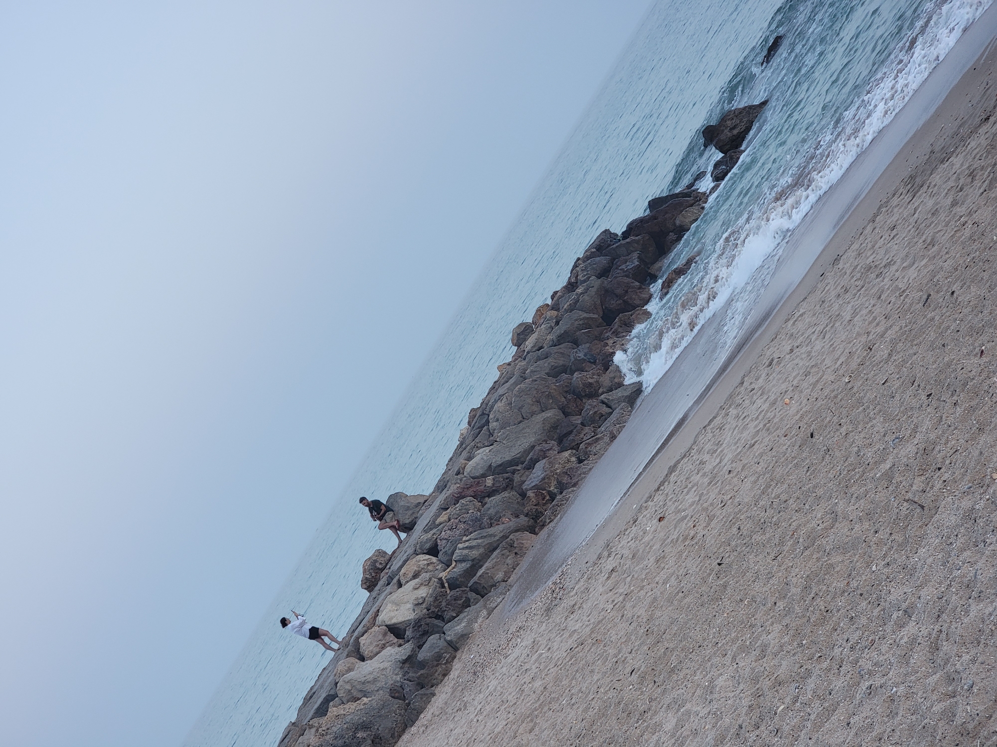 In Quarteira, Portugal, a person dressed in white and black shorts stands on a rocky breakwater, holding a phone and appearing to take a photo. Another person, dressed in a black t-shirt and shorts, is seated on a large rock further down the breakwater, looking towards the water. The breakwater is composed of large, rough stones and slopes down towards a sandy beach where small waves are breaking. The ocean stretches out to a hazy horizon, and the sky is overcast and light blue. The scene is captured from a low angle, with the breakwater and beach diagonally bisecting the frame.