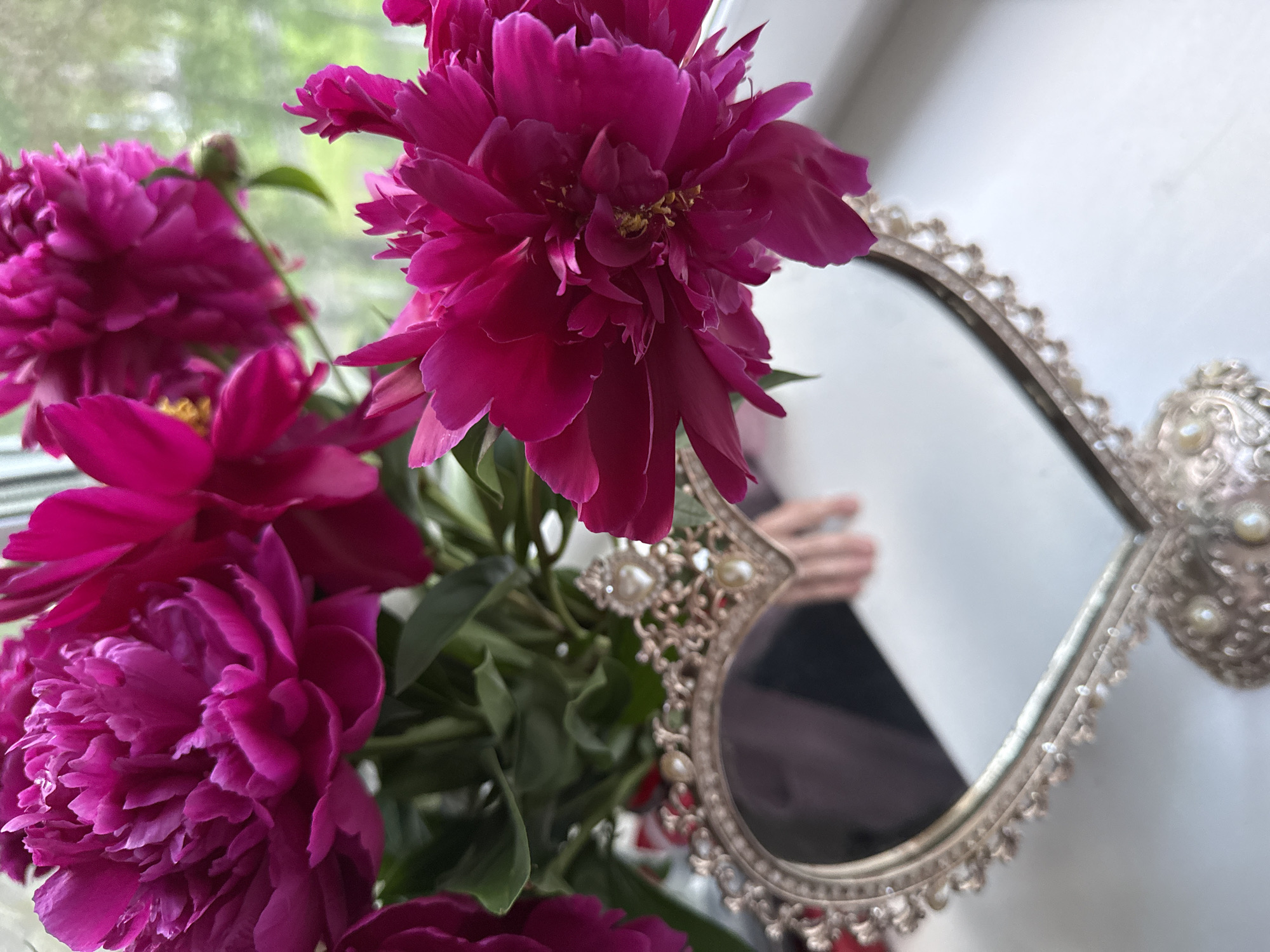A close-up, vertical shot features several bright pink peonies and a heart-shaped decorative mirror. The mirror has an ornate, antique-style frame with pearl accents and is positioned in the foreground. The reflection in the mirror shows a blurred hand with rings, suggesting a person is holding or interacting with the scene. The peonies are the dominant subject, with some in sharp focus and others slightly blurred due to shallow depth of field. The background is out of focus, showing a hint of greenery through what appears to be a window. The overall lighting is soft and natural. The scene is indoors, on a white surface, possibly a windowsill.