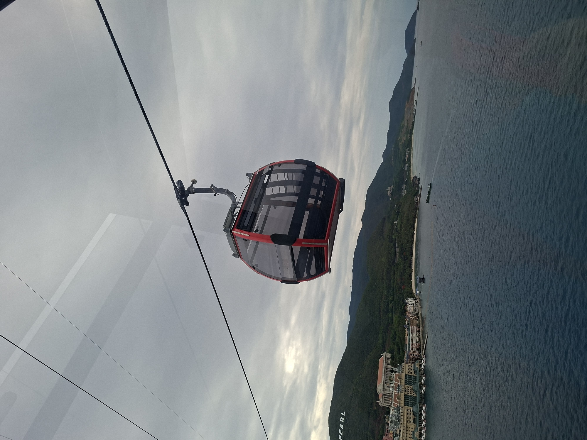 An aerial view looking out from inside a cable car over Nha Trang Ward, Vietnam. The cable car is predominantly red and black with transparent windows, suspended by thick black cables extending diagonally. Outside, the landscape features a mountainous coastline covered in lush greenery. Alongside the coast, a line of buildings is visible, suggesting a densely populated area. The sea stretches out beyond the coastline, its color ranging from deep blue to lighter shades near the shore. The sky above is overcast with varying shades of gray clouds.
