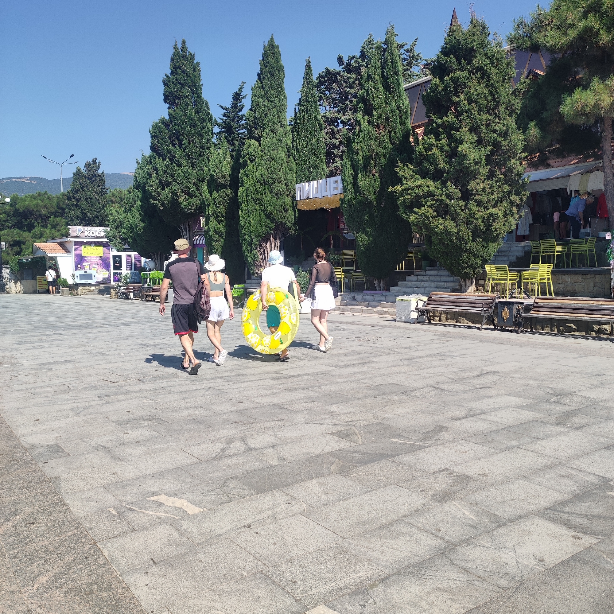 The image shows a paved walkway on a sunny day. Four people are walking away from the camera. The person in the front left is a male wearing a dark t-shirt and black shorts. The person next to him is a female wearing a white hat, a white top, and white shorts. They are both wearing sandals. The third person is a male wearing a blue hat, a white t-shirt, and teal shorts. He is carrying a yellow inflatable ring with lemon patterns. The last person is a female wearing a black sheer top and a white skirt. Behind them, there is a building with a sign that reads "ПИЩЕБЛОК" in large letters. The building has an awning and tables and chairs outside. There are also tall green trees lining the walkway. In the background, there are hills and a clear blue sky.