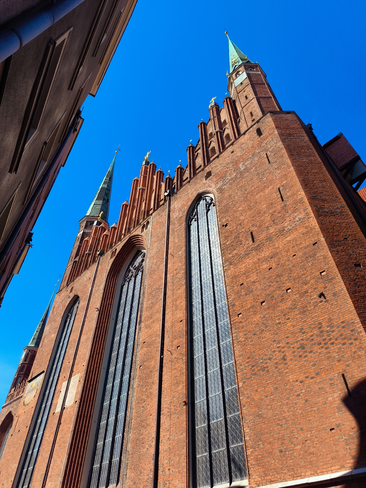 The image captures a low-angle view of the St. Mary's Church (Bazylika Mariacka) in Gdańsk, Poland, under a clear blue sky. The massive brick structure of the church dominates the frame, with its characteristic Gothic architecture. Tall, arched windows with intricate leaded glass are visible along the side of the building. The church features multiple spires, one of which, covered in green copper, is prominent in the upper portion of the image, topped with a golden finial. The textured red brickwork, interspersed with darker bricks and small square indentations, shows the age and construction of the building. Adjacent to the church, on the left side of the frame, is the facade of a tall, narrow building, also constructed of brick, with a darker, smoother finish. The perspective emphasizes the grandeur and height of the church. The lighting suggests it is a sunny day, with shadows cast on the brickwork.