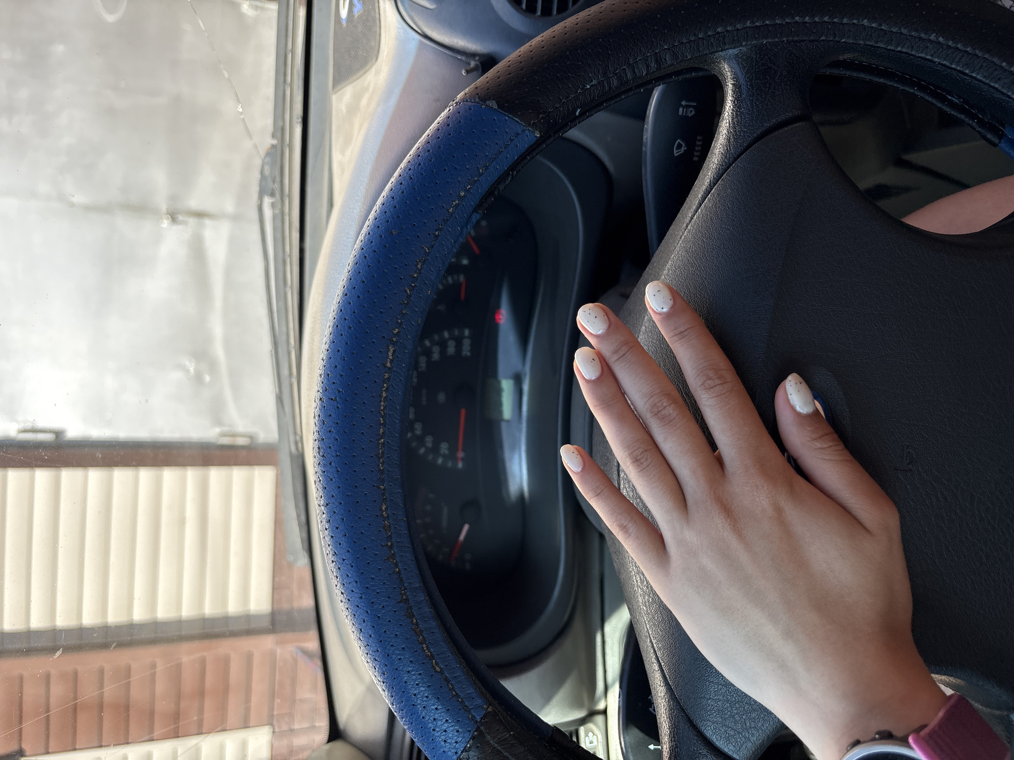 The image shows a close-up view of a person's hand with white nail polish and speckles resting on the steering wheel of a car. The steering wheel has a blue cover. The dashboard with a speedometer is visible behind the steering wheel. The windshield shows a reflection of a building with horizontal siding and some sky. The scene appears to be outdoors, possibly in a parking area. The location is specified as Прикамский, Russia.