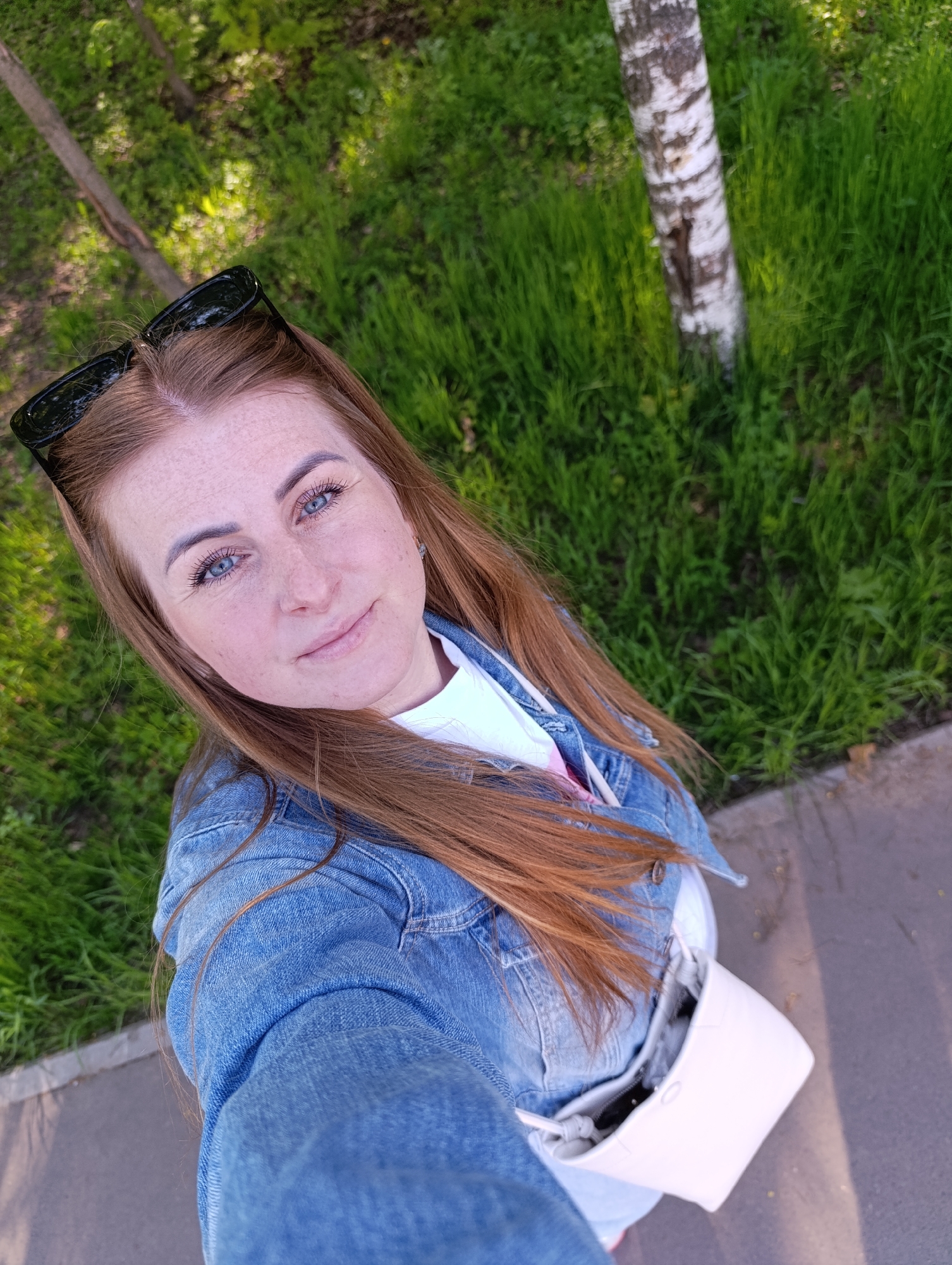 A fair-skinned woman with freckles, long red hair, and blue eyes is positioned in the foreground of the image, appearing to take a selfie. She wears sunglasses perched on her head and is clad in a blue denim jacket over a white shirt. A light-colored shoulder bag hangs by her side. The background consists of green grass and the trunk of a birch tree. The setting appears to be a park within an unidentified city. A section of sidewalk is visible at the bottom right.