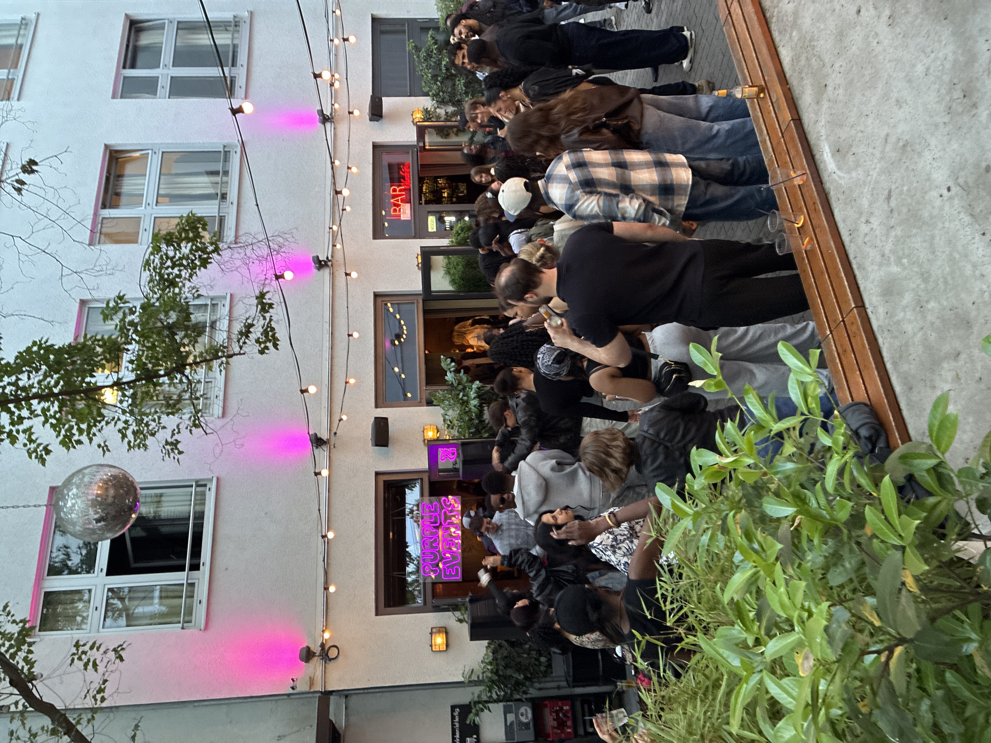 This image depicts an outdoor courtyard of a venue in Berlin, Germany. The venue appears to be a bar or club, with a neon sign reading "PURPLE EVENTS" above an entrance, and another sign saying "BAR" in red neon above a separate entrance. The courtyard is populated by a diverse group of people, dressed in casual attire, suggesting a social gathering or event. String lights are strung across the courtyard, and pink uplighting illuminates the facade of the building. A disco ball is suspended from the ceiling, contributing to the festive atmosphere. Some individuals are standing in groups, while others are in the process of ordering drinks at the bar or enjoying beverages. The overall scene conveys a lively and engaged crowd enjoying a night out in an urban setting.