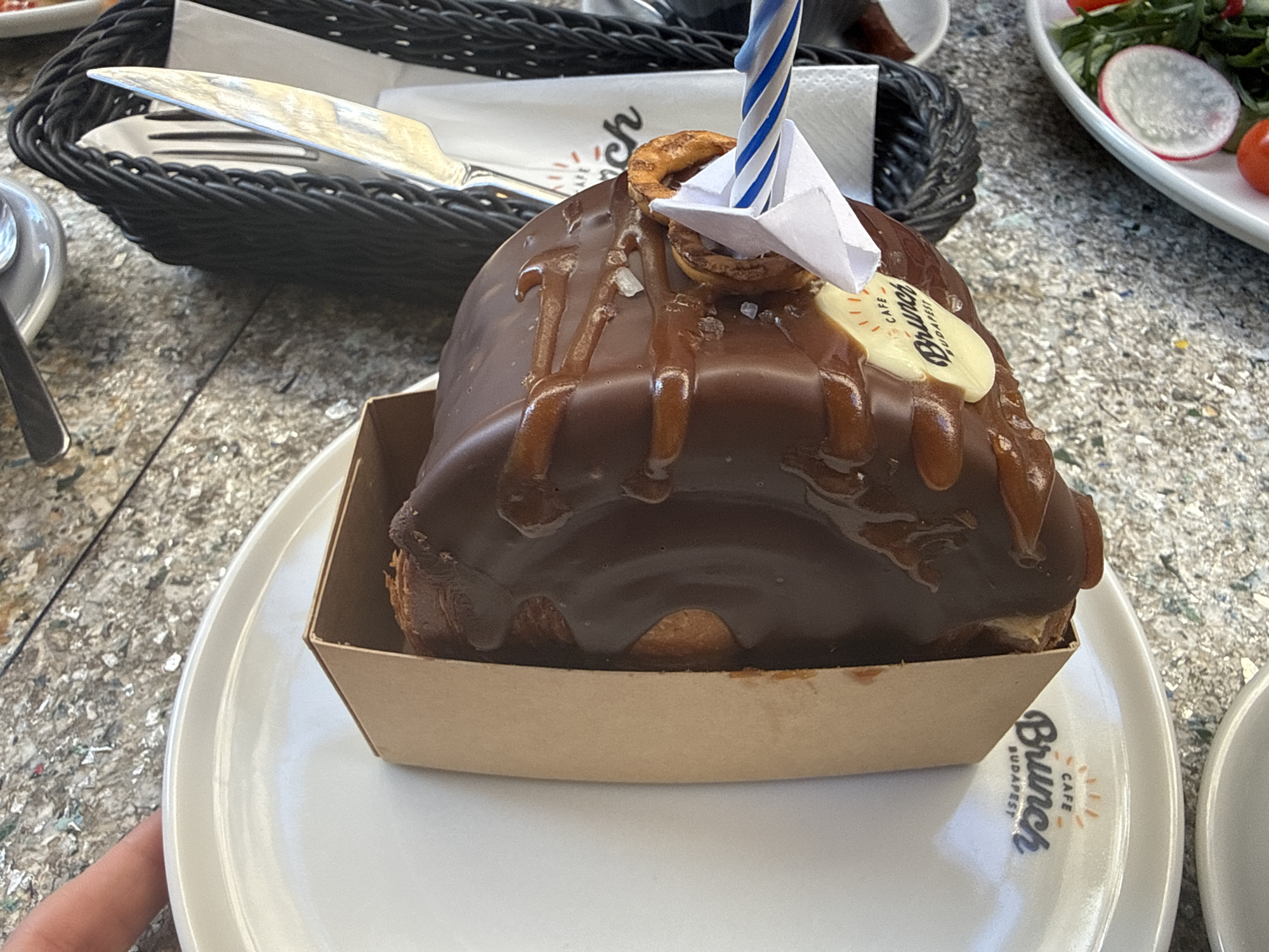The image presents a close-up view of a decadent chocolate-covered pastry, likely a large croissant or cake, set on a white ceramic plate. The pastry is generously coated in dark chocolate icing with visible caramel drizzles and sprinkles of sea salt, nestled in a light-brown cardboard tray.
It is adorned with festive elements: a miniature white paper boat with blue stripes, a blue and white striped birthday candle, and a small, golden-brown pretzel-like decoration. A white, oval plaque, possibly made of chocolate, is embedded in the icing, displaying the text "CAFE Brunch Budapest" accompanied by a sun graphic. This same "CAFE Brunch Budapest" logo is also printed on the rim of the white ceramic plate, clearly indicating the location to be Budapest, Hungary, at a cafe called Brunch.
In the background, a black woven basket containing a knife and fork is visible. To the right, a white plate holding a fresh salad with green leaves, sliced radishes, and cherry tomatoes suggests a meal is taking place. The table surface is a mottled, light grey texture. A person's thumb is visible at the bottom left, holding the plate. The bright lighting indicates it is daytime. The candle and celebratory decorations suggest a special occasion, such as a birthday or a festive treat.