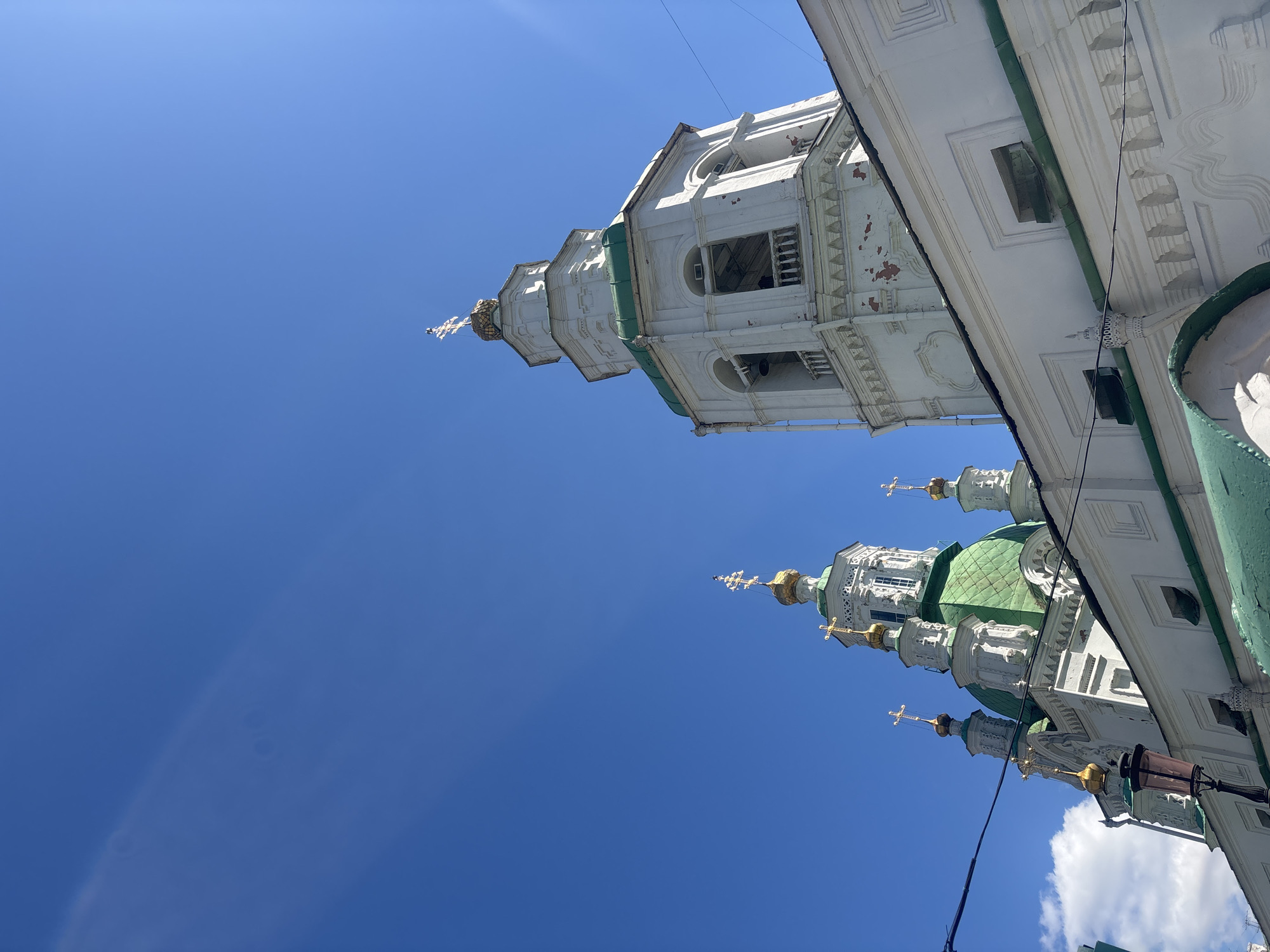 A low-angle, upward shot captures a portion of the Holy Intercession Cathedral in Krasnoyarsk, Russia. The church's white facade is accented with green roofing on the domes and trim. The blue sky provides a contrasting backdrop. A section of the building's architecture is also visible in the right upper corner.