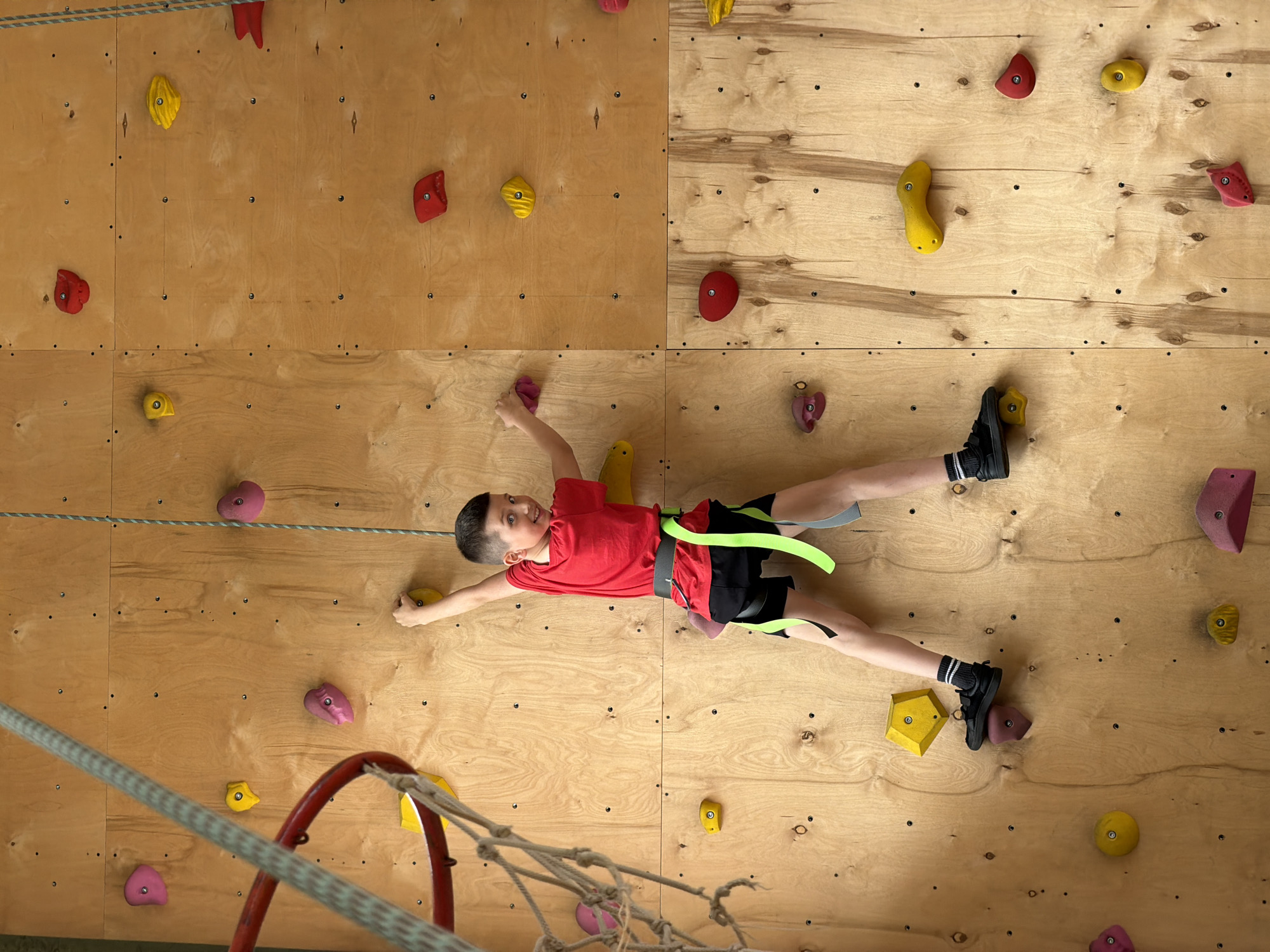 A boy is rock climbing indoors on a wooden wall with colorful handholds and footholds. He is wearing a red t-shirt, black shorts with green accents, and climbing shoes. He is using a climbing harness with neon green straps. The wall is made of light-colored wood panels joined together. A climbing rope is visible in the lower left corner of the frame. The photo was taken from a high-angle perspective. The location is in Izhevsk, Russia.