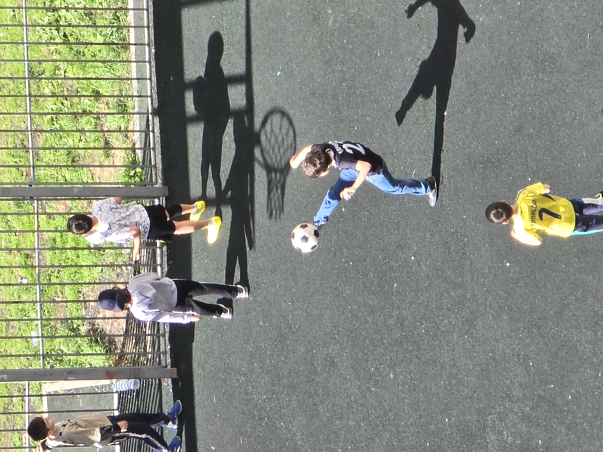 The image is a high-angle view of a sports court, possibly in the city of Khimki, Russia. On the left side of the frame, four children stand near a metal fence that separates the court from a grassy area. One child wears a beige jacket and dark pants with blue shoes. The second child wears a grey sweatshirt, dark pants, and a baseball cap. The third child wears a white shirt and dark shorts with yellow shoes. The fourth child is partially obscured.
On the right side of the frame, three other children are engaged in playing. One child, wearing a black jersey, jeans, and sneakers, kicks a soccer ball. Another child, wearing a yellow soccer jersey with the number 7, lies on the ground near the edge of the frame. There are several prominent shadows on the court, including one that appears to be of a basketball hoop. The court surface is a dark gray color.