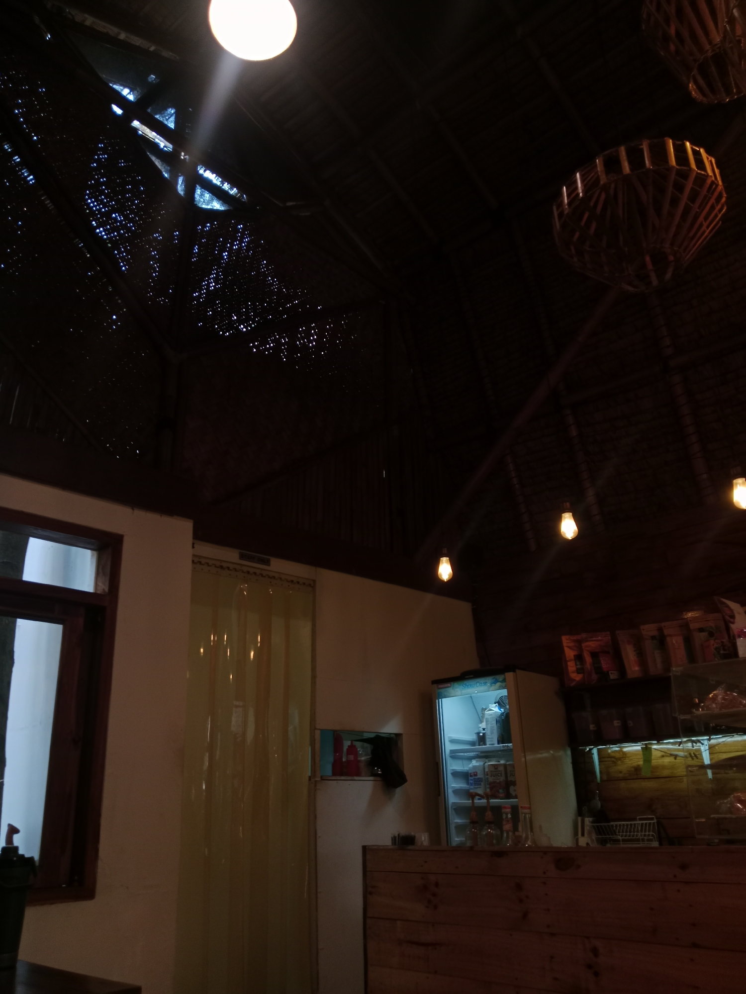 The image depicts the interior of a dimly lit establishment, possibly a cafe or a shop, with a thatched roof. The ceiling features exposed wooden beams and is partially covered by a woven material that allows some light to filter through. Two large, basket-like light fixtures hang from the ceiling. A bright circular light source is visible in the upper left corner, casting a strong beam.
In the foreground, a wooden counter runs horizontally across the bottom of the frame. Behind the counter, a white refrigerator with its door ajar reveals bottles and cartons inside. Above the refrigerator, shelves are stocked with various packaged goods. To the left of the refrigerator is a window with a dark wooden frame. To the left of the window, a dark green object, possibly a bucket or planter, is visible on a dark surface.
To the right of the window, a light-colored, translucent curtain hangs from a rod, obscuring what lies beyond. A framed picture is hung on the wall to the right of the curtain. Several pendant lights with exposed bulbs hang from the ceiling in the middle ground. The overall ambiance is rustic and warm, with the limited lighting creating a sense of intimacy. The specific location in Bandung City, Indonesia, is not identifiable from the visual information provided.