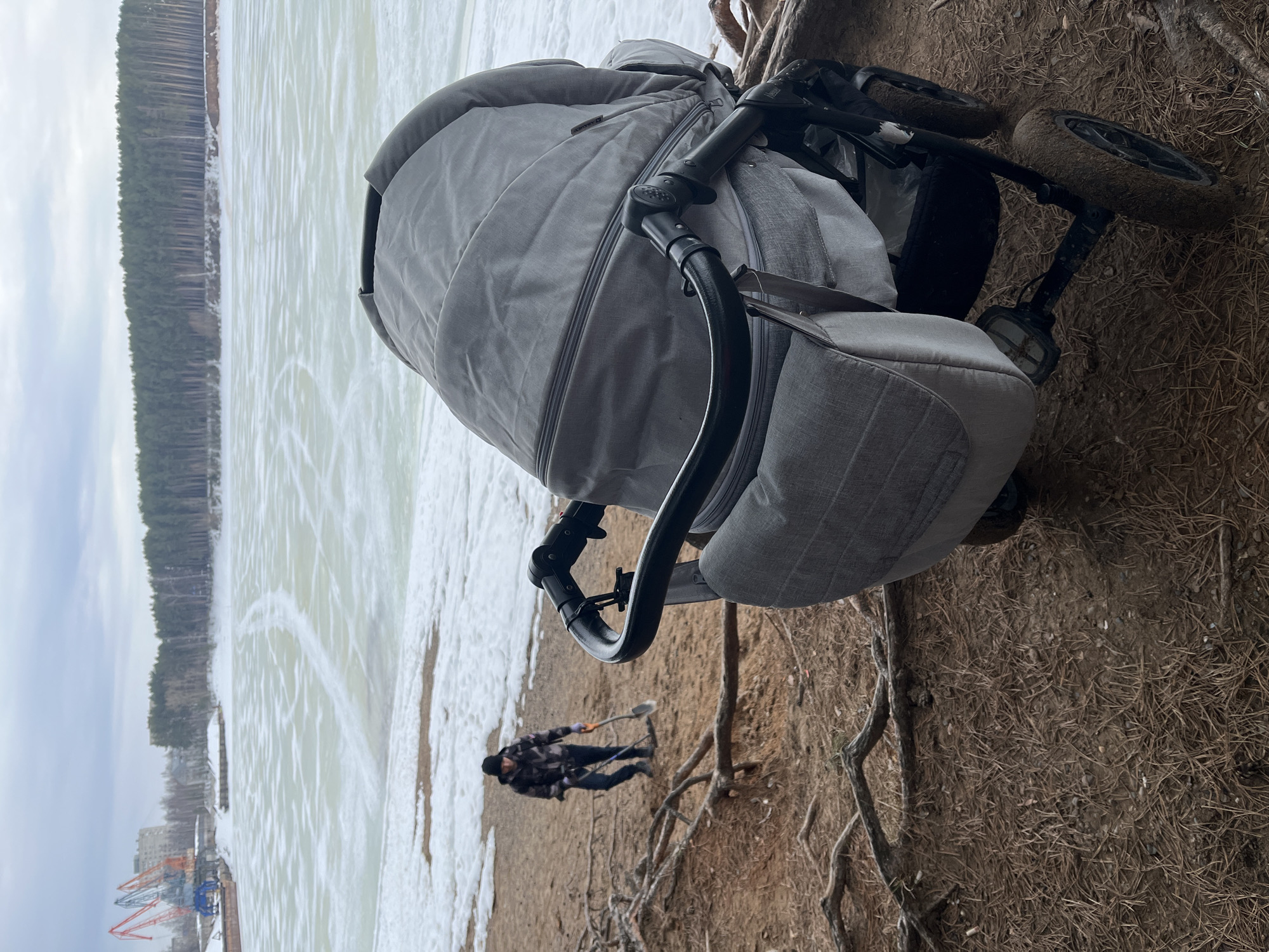 The image shows a gray baby stroller positioned on a dirt and root-covered ground. In the background, there is a body of water covered with ice and snow, with trees lining the opposite shore. In the lower center of the frame, a person is visible holding a shovel. In the lower left corner, there are structures that appear to be cranes and buildings in the city of Chaikovsky, Russia.