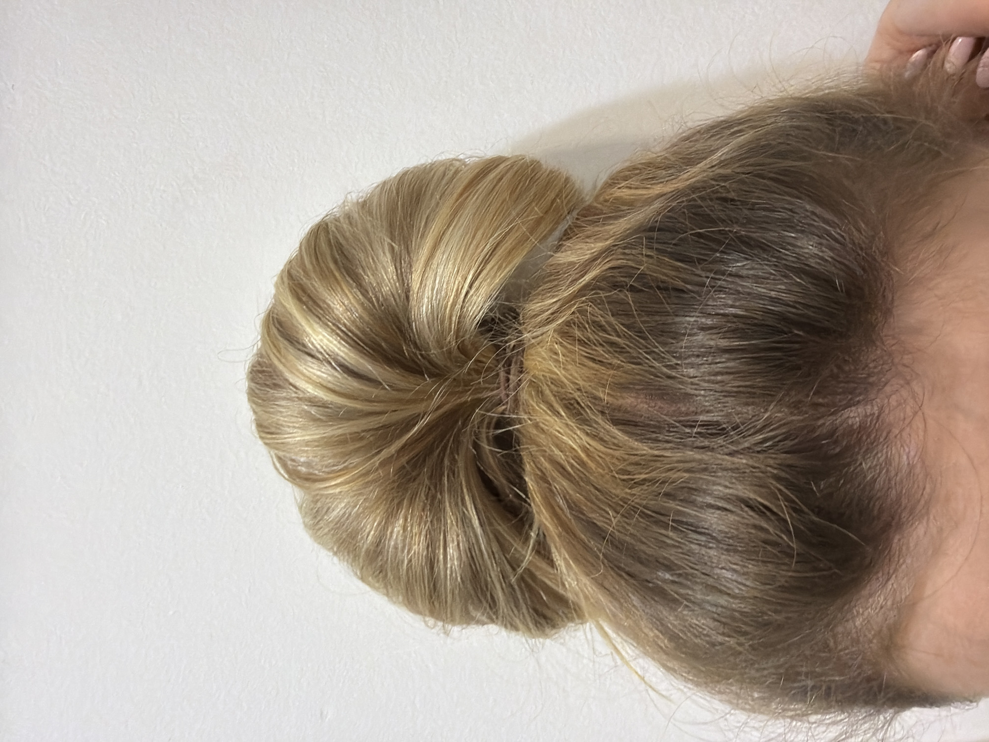 A close-up, eye-level shot depicts a person's blonde hair styled in a bun. The bun is centrally located in the frame, revealing the detailed texture of the hair. The background is a plain white surface. A portion of the person's skin, including part of their hand, is visible in the top right corner. No location is identifiable from the image.