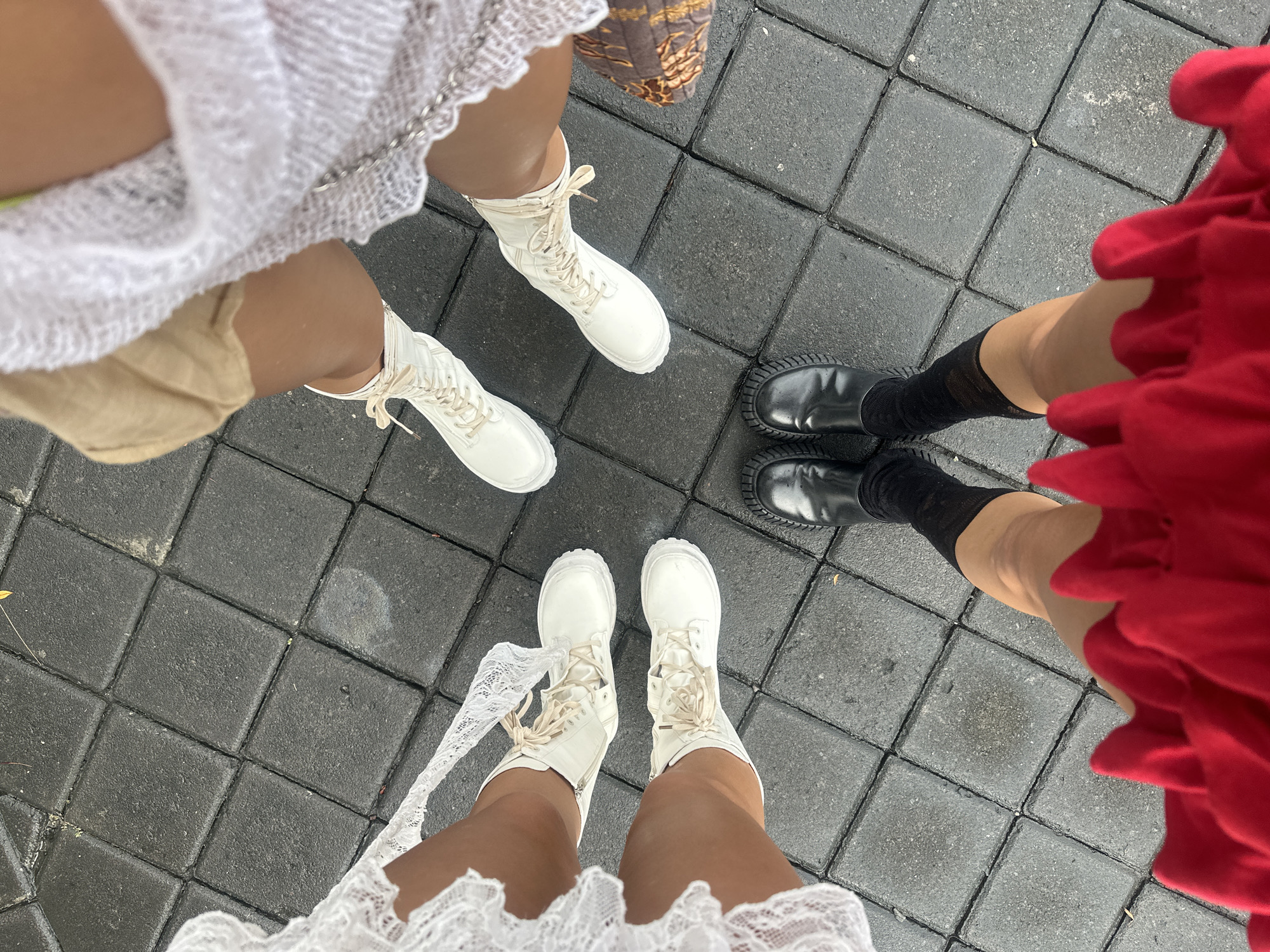 Four individuals stand on a tiled surface in North Kuta, Indonesia. Three are wearing white boots with tied laces, and one wears black shoes with black socks. Two individuals wear white, frilly dresses. The third wears tan shorts with a white dress, and the fourth wears a red dress. A brown purse is also visible.