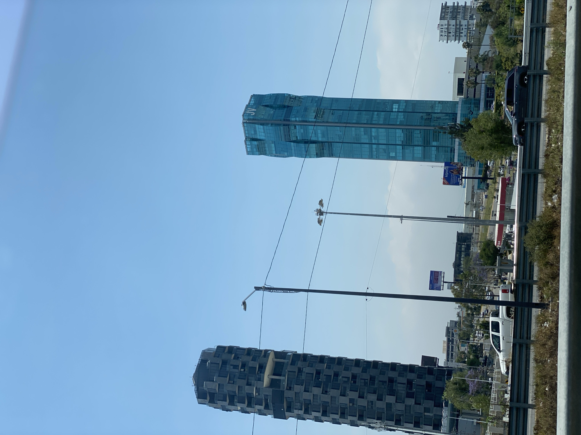 The image shows two tall buildings in an urban environment with a bright blue sky. The building on the left is shorter and darker, with a rectangular shape and a facade of repeated square modules. The building on the right is taller and blue, with a more modern and undulating design, and is branded with "JV" on top. Power lines intersect the view. A highway runs in front of the buildings, with cars and a guardrail visible. Street lights are also apparent along the road. In the background, there are other buildings and greenery. The location is the city of Unknown, Unknown.