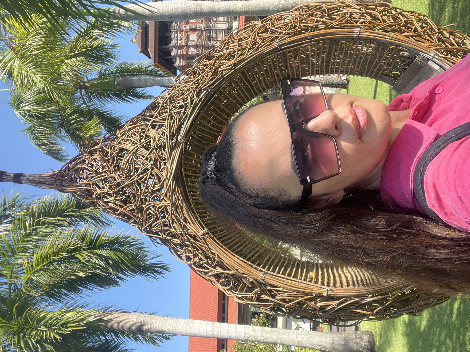In Kuta Selatan, Indonesia, a woman with brown hair pulled into a high ponytail, wearing large sunglasses and a vibrant pink collared shirt, poses for a selfie. She is seated within a large, woven rattan chair, which resembles a bird's nest. The background features lush green grass, several tall palm trees with their fronds reaching towards a clear blue sky, and traditional Balinese architecture with red roofs and intricate carvings. The scene is bathed in bright sunlight, casting shadows on the ground.