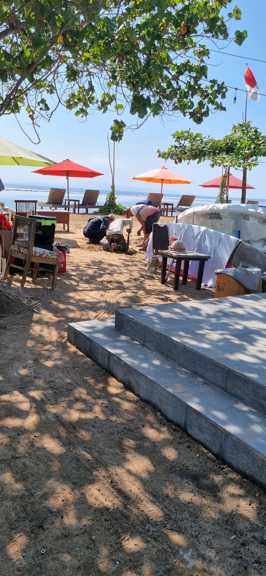 This image captures a scene on a beach in Denpasar, Indonesia, likely a section of the coastline known for its resorts and tourist amenities. The foreground features a sandy area with some newly laid grey stone steps. The middle ground displays a collection of beach furniture including lounge chairs and umbrellas in shades of green, orange, and red. Several people are present, with two individuals actively engaged in digging or working with the sand, one crouching and holding a shovel. Another person is visible behind them, partially obscured. In the background, the ocean is calm, with a hazy horizon suggesting a clear, sunny day. A red and white flag, possibly the Indonesian flag, is flying in the distance. The overall impression is one of a beach preparing for or undergoing some activity, with a mix of leisure items and construction elements.