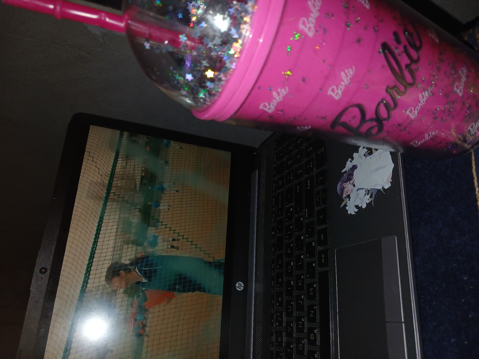 The image shows a laptop partially obscured by a pink Barbie-themed cup with a straw and glitter inside. The laptop screen displays a person playing a sport within a net-enclosed area. The keyboard of the laptop is visible, with a sticker of a woman on the right side. An HP logo is visible on the laptop's body. A dark blue surface is partially seen in the bottom right corner of the image. There is no information to determine the location in the city of Salar, Uzbekistan.