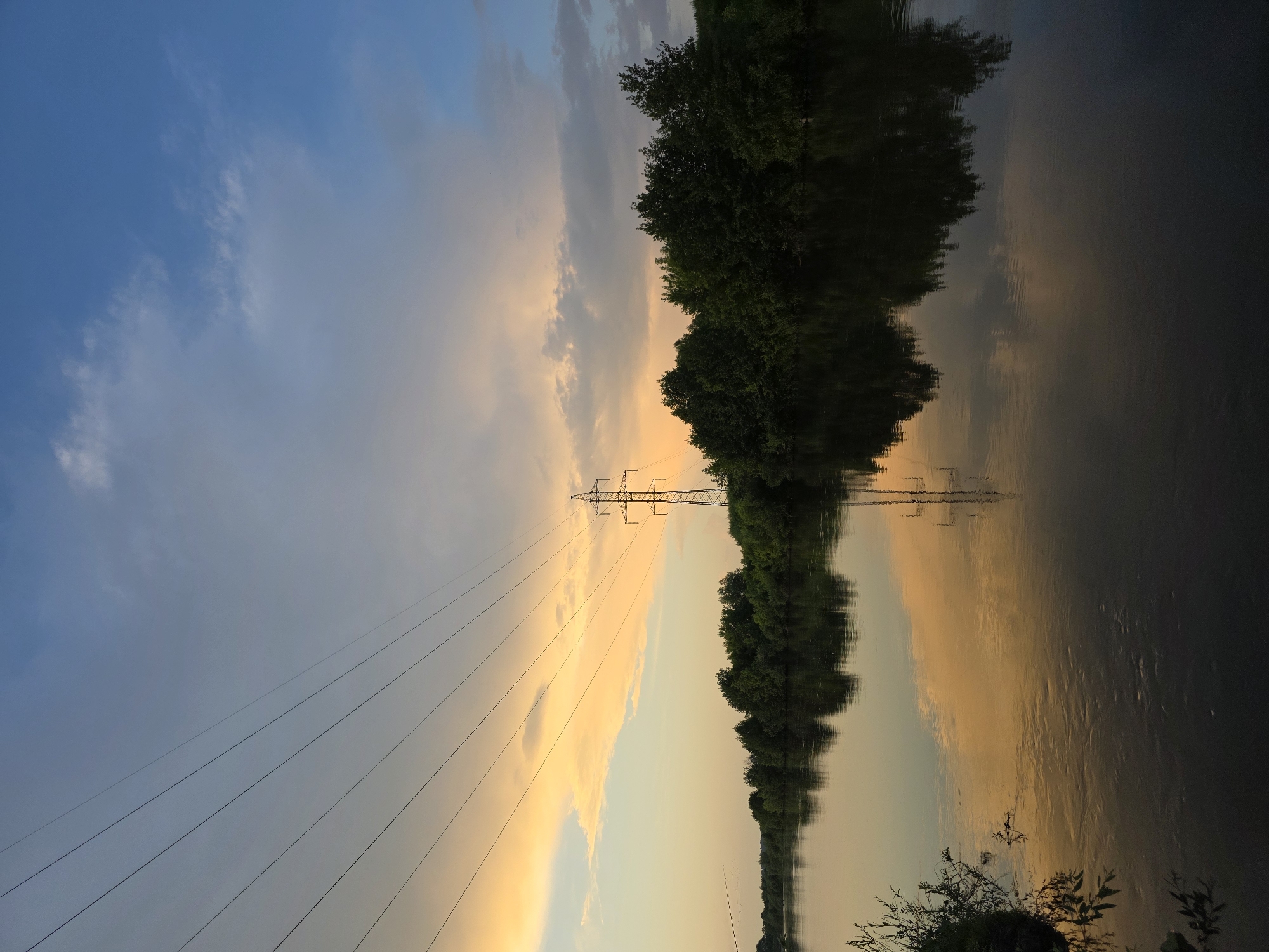 The image depicts a serene sunset over a wide river in the city of Togashevo, Russia. The sky is a gradient of colors, transitioning from a pale yellow and orange on the horizon to a soft blue overhead, with scattered clouds. Several power lines stretch diagonally across the sky, leading the eye towards a tall electricity pylon situated on the far bank. The river's surface is calm, perfectly reflecting the sky, the pylon, and the dense line of green trees that borders the opposite bank. On the left bank, some vegetation and branches are visible close to the water's edge. The overall atmosphere is peaceful and tranquil, characteristic of a natural landscape at dusk. There are no people or animals visible in the scene.